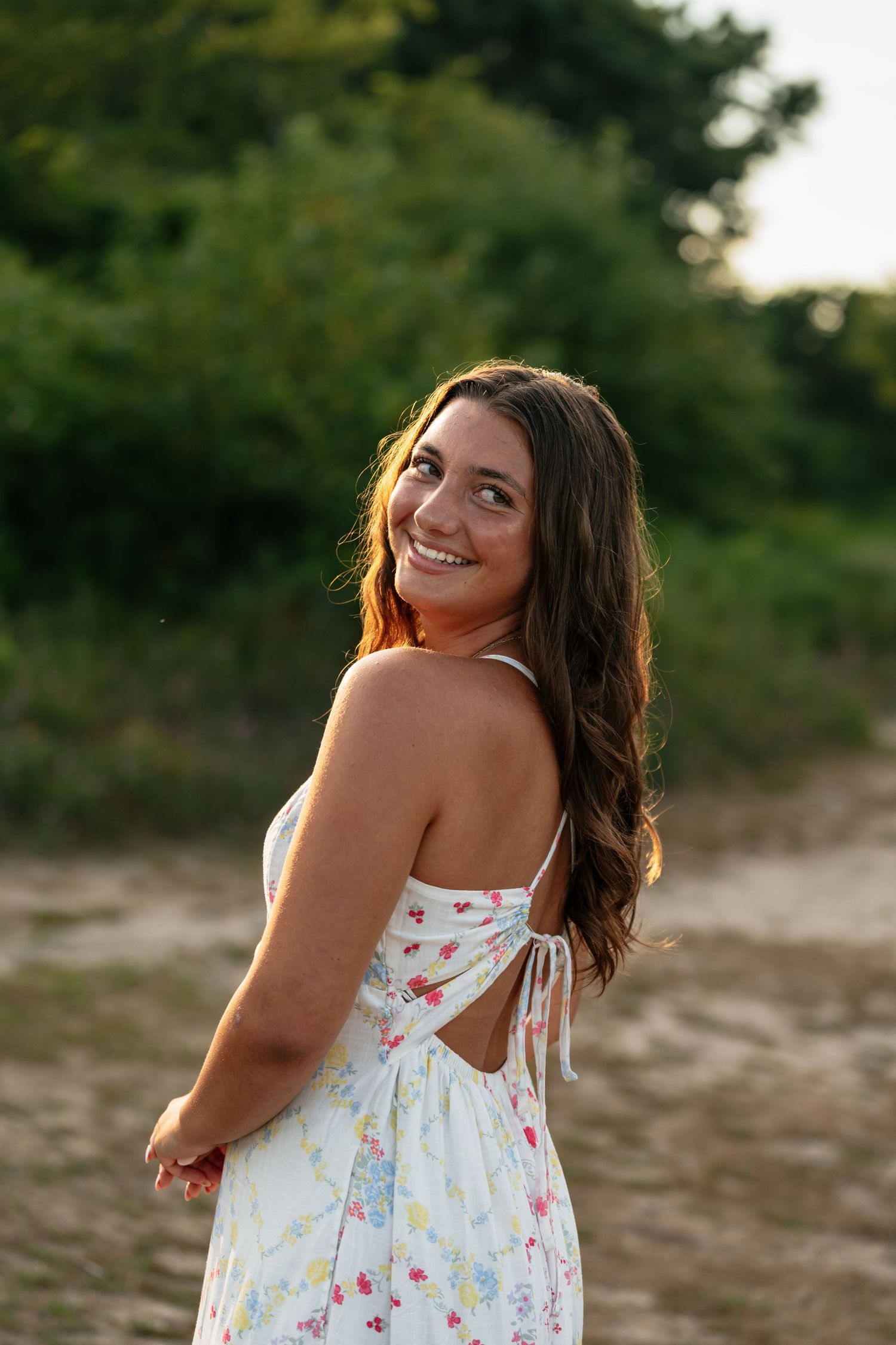 Person in white floral dress poses playfully during sunset photoshoot.
