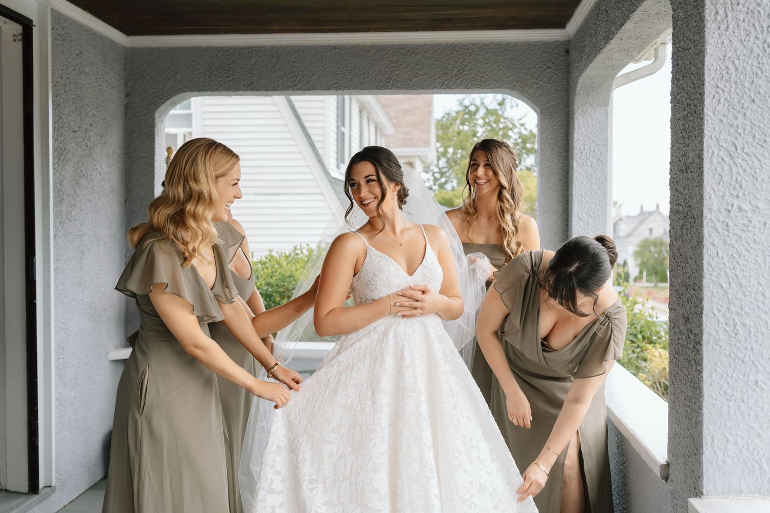 Bridesmaids in sage green dresses help adjust a wedding gown on a covered porch.