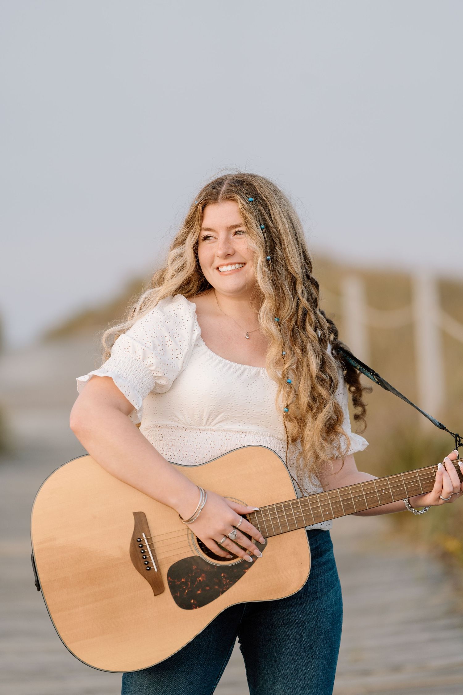 Musician playing acoustic guitar outdoors wearing white top and jeans.