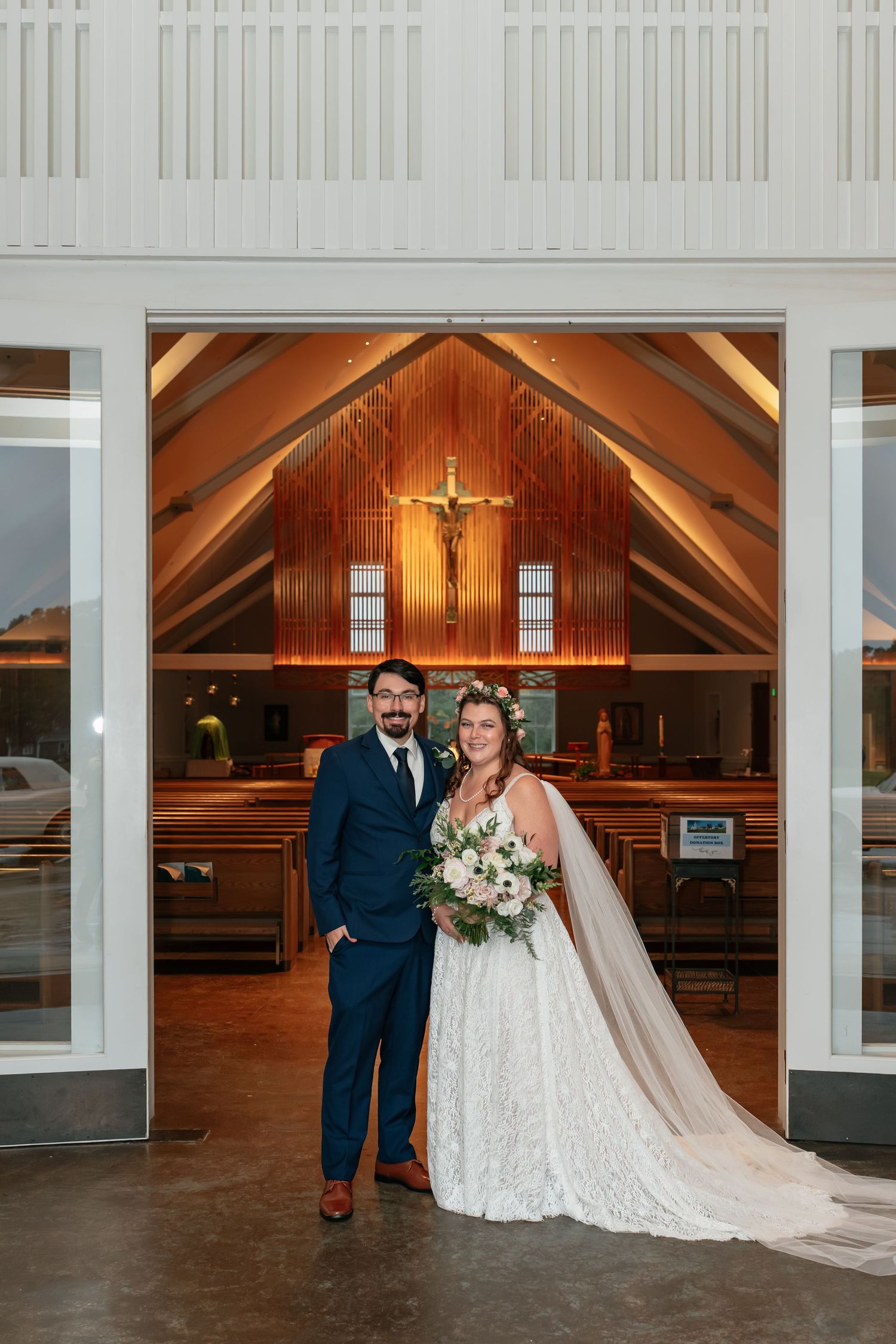 Wedding couple poses in doorway of church with wooden crucifix visible in background.
