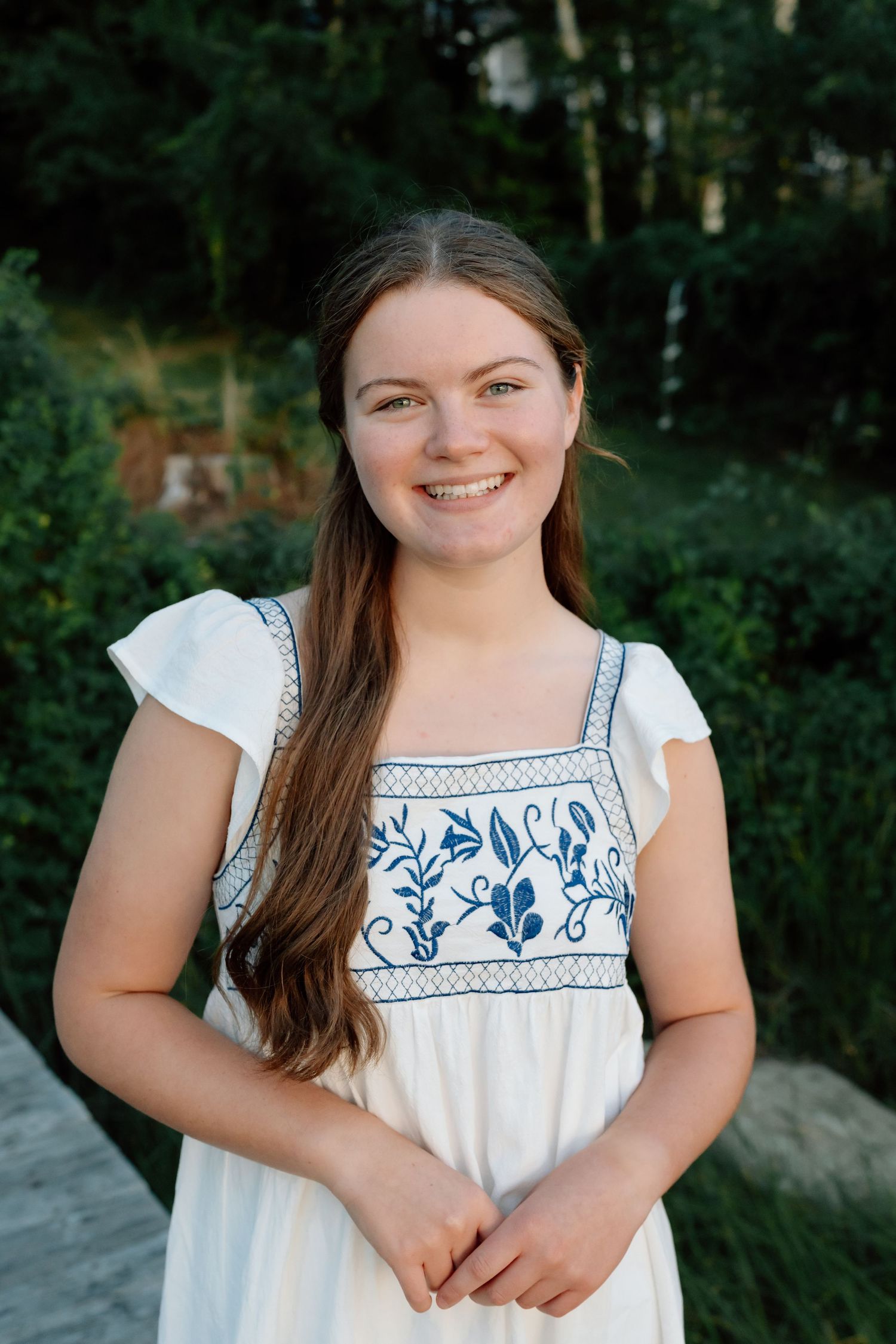 Summer portrait in white embroidered dress against lush green forest background.