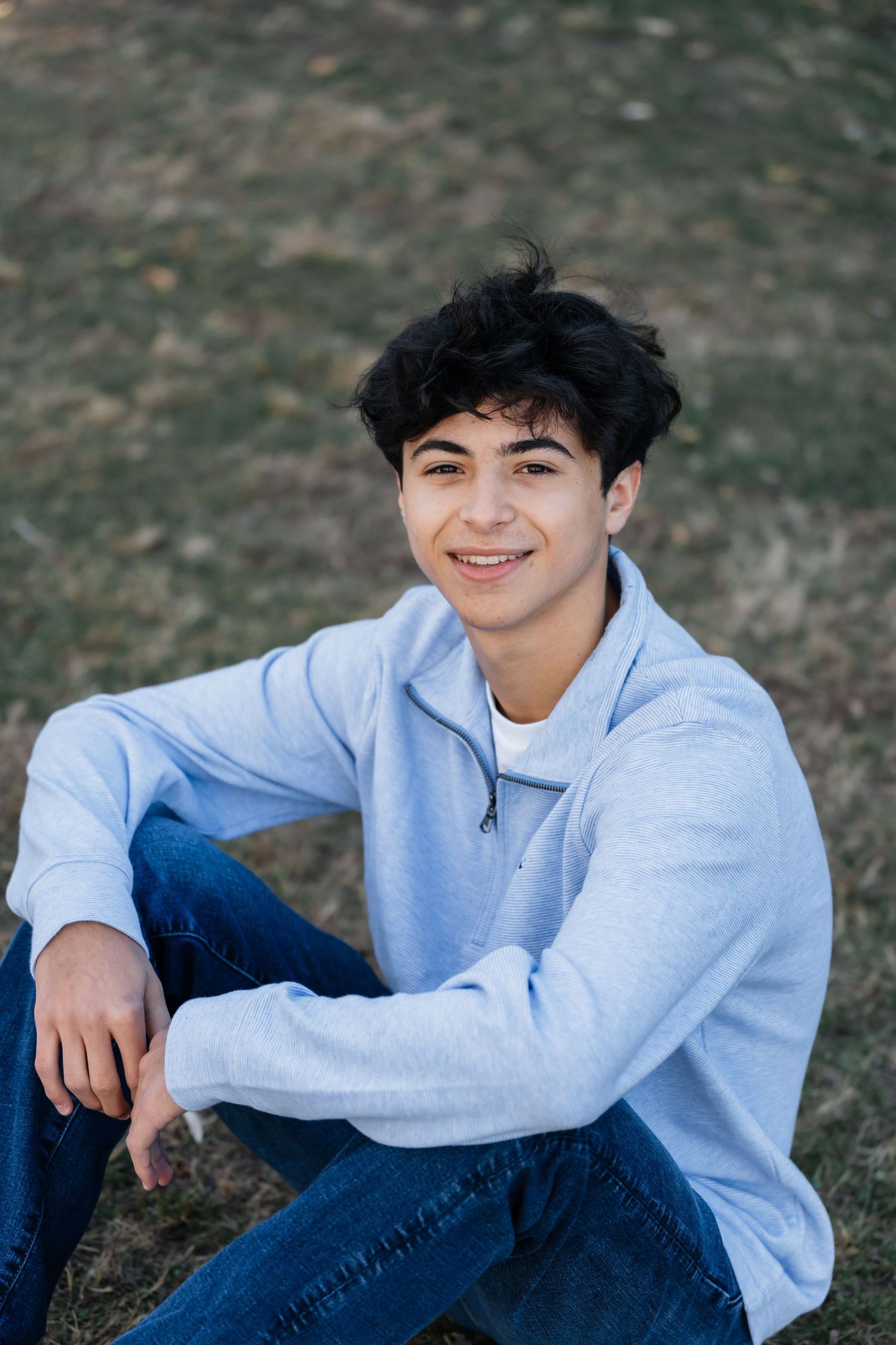 Person in light blue sweater and jeans sits casually on grass during an outdoor photoshoot.