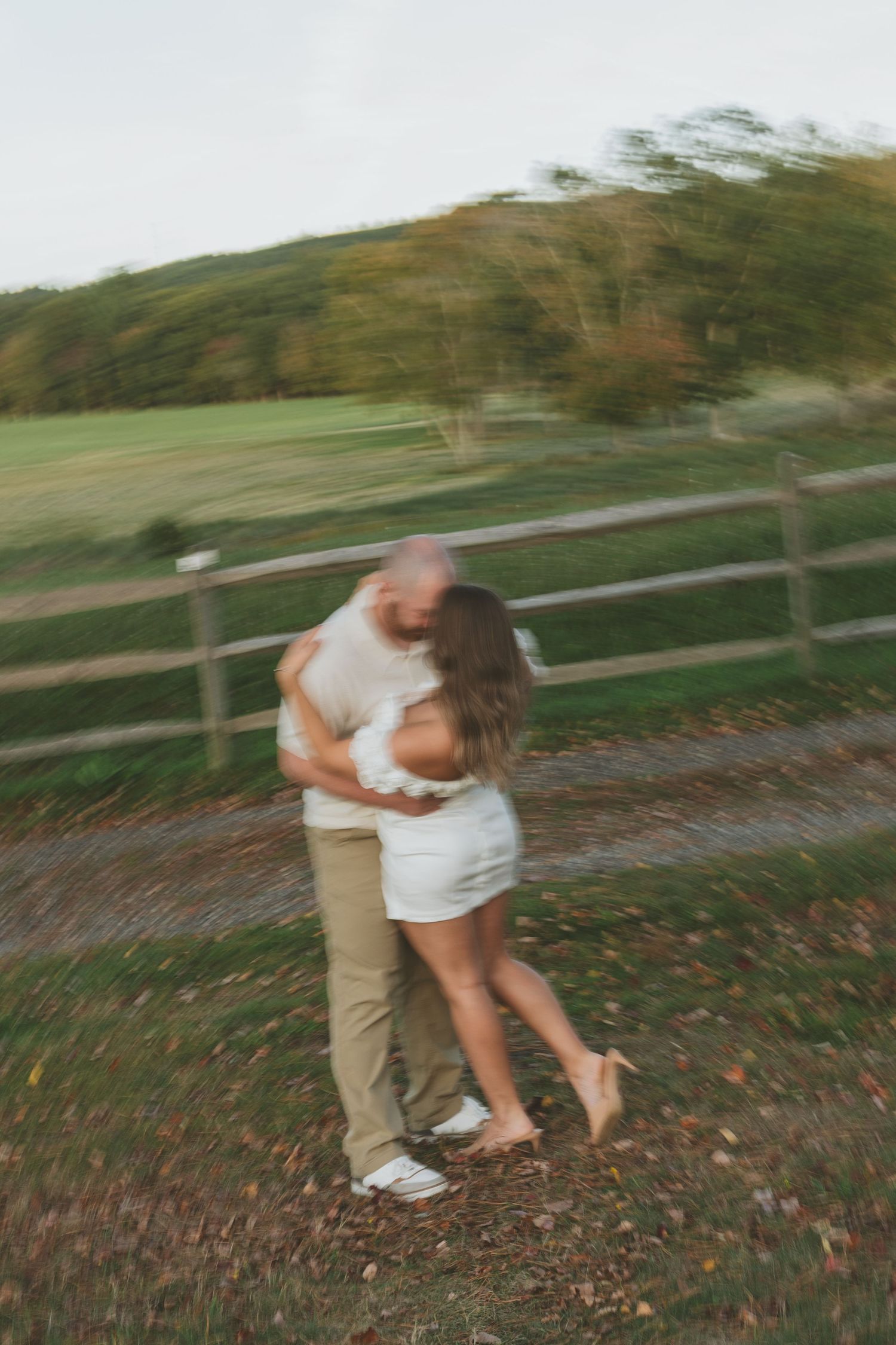 Motion-blurred photo of couple dancing on leaf-covered ground by wooden fence.