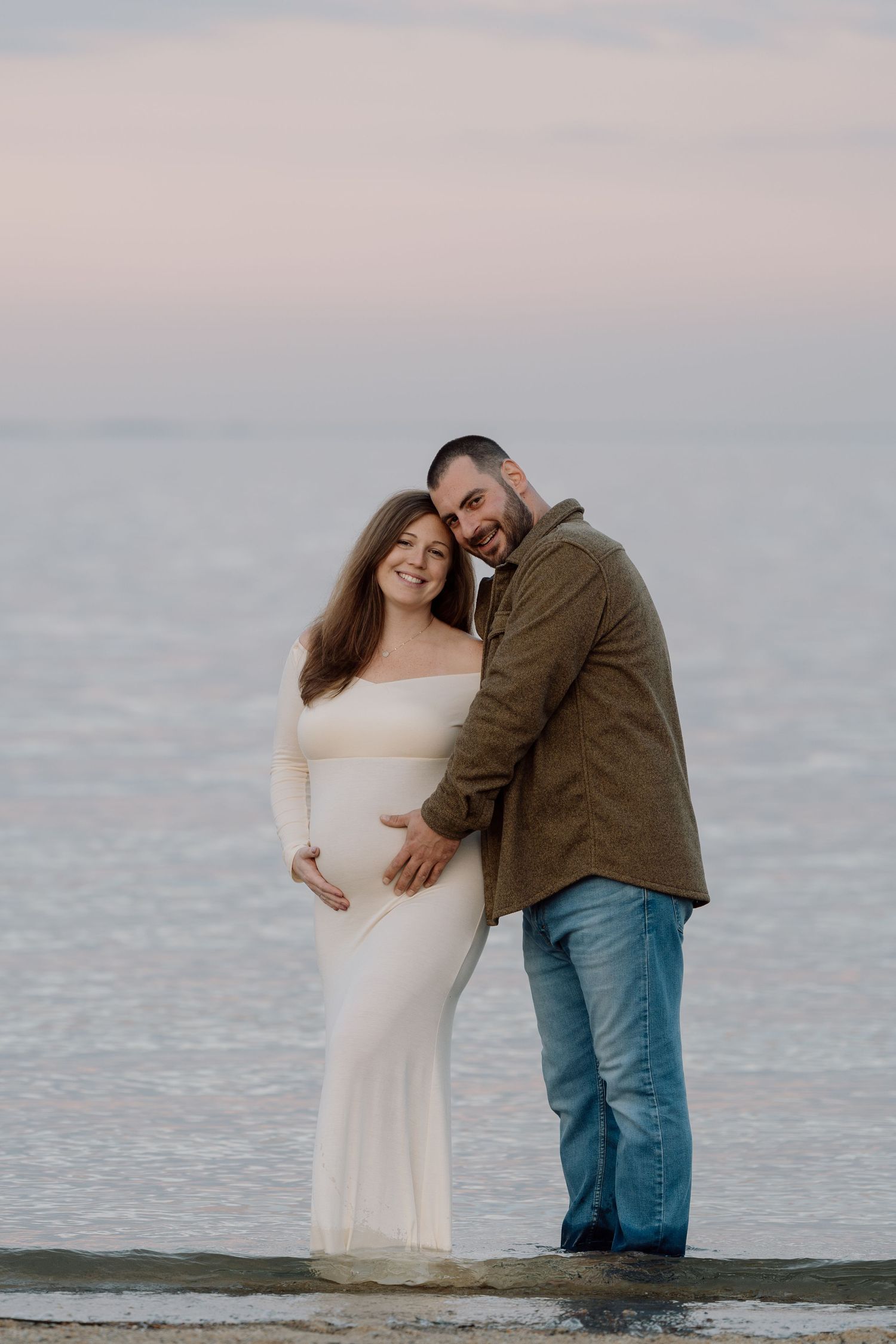 A couple embraces on the beach at sunset during a maternity photoshoot.