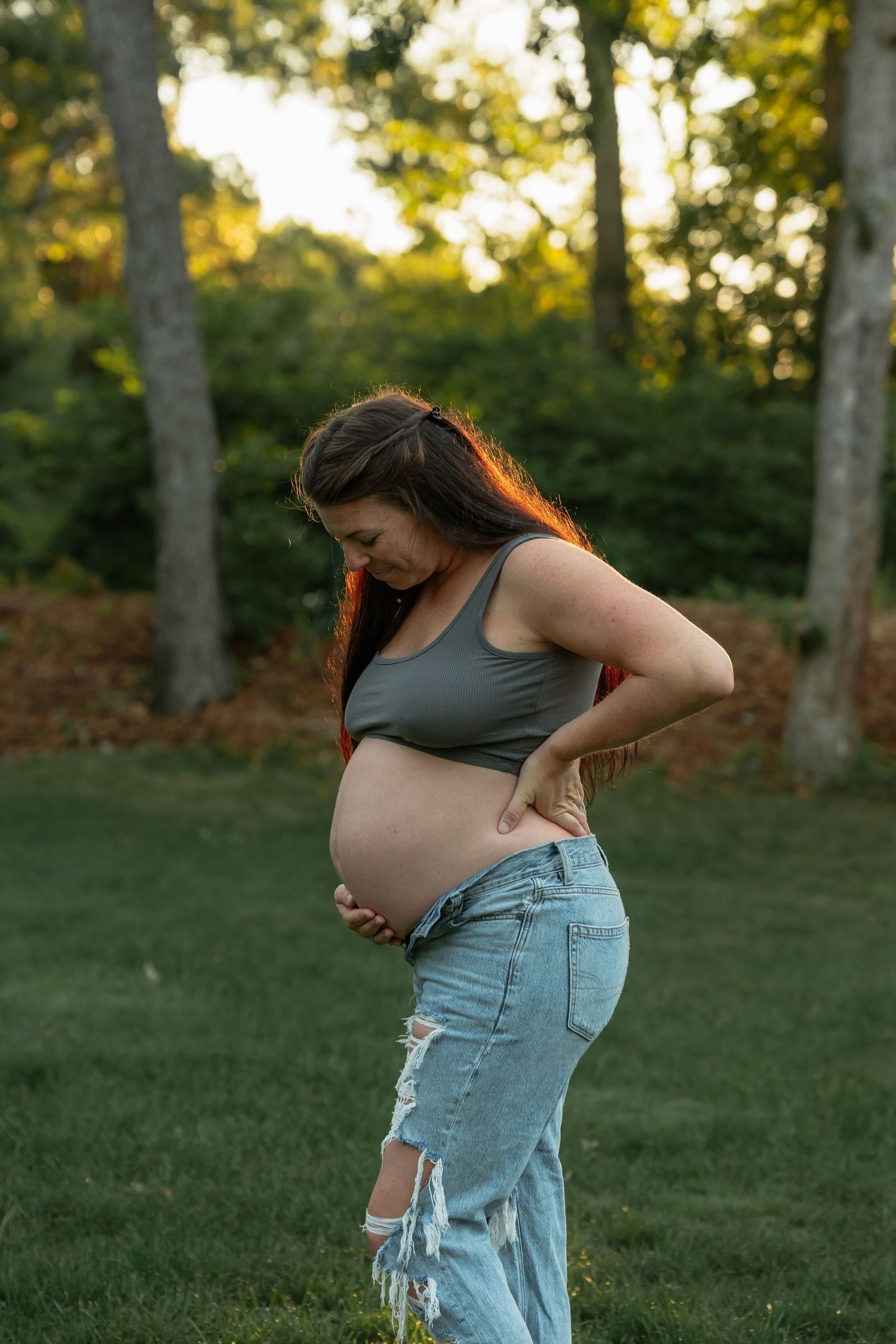 Expectant mother in ripped jeans poses against green trees during golden hour.
