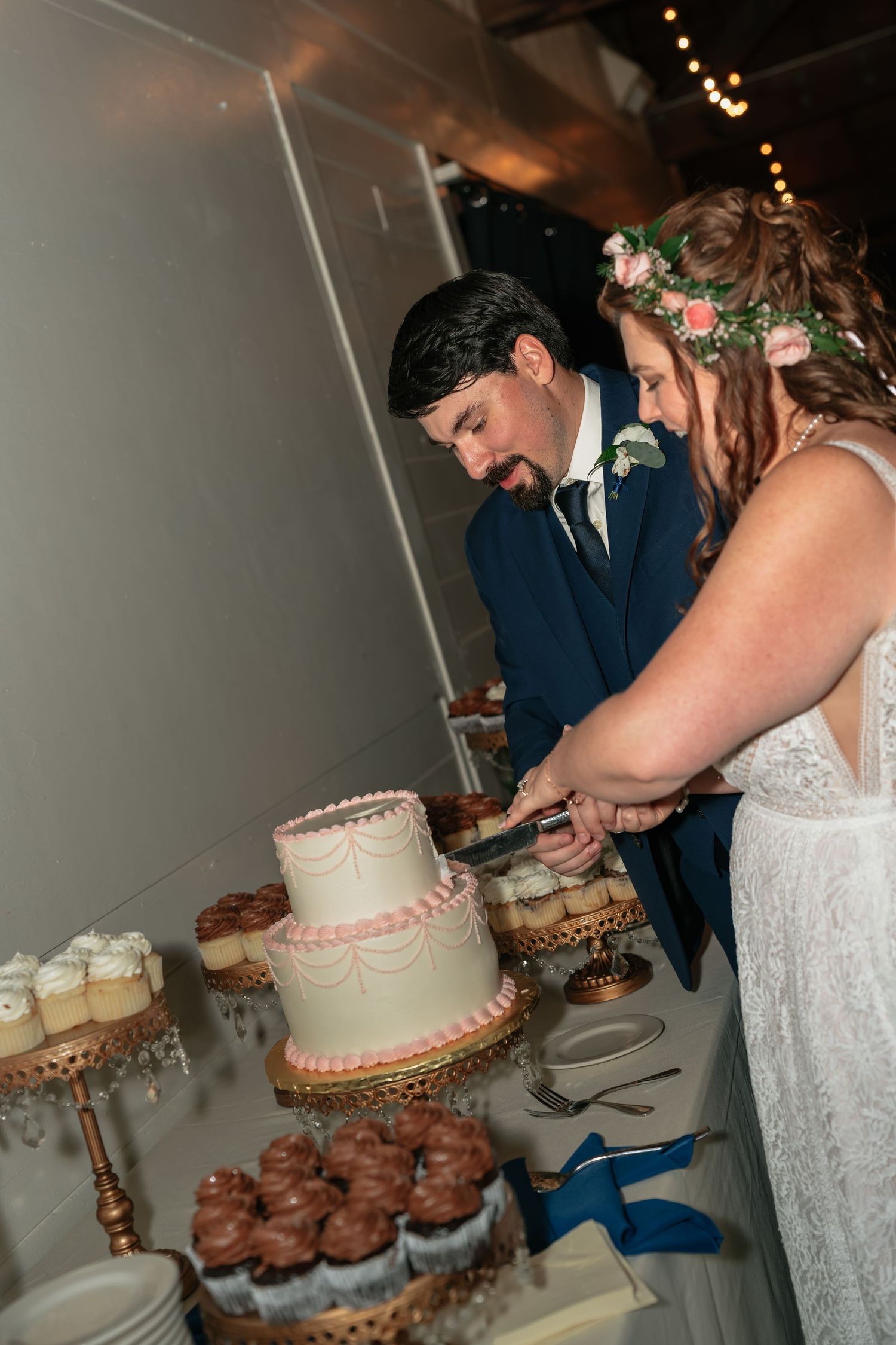 Couple cuts their wedding cake together at reception dessert table.