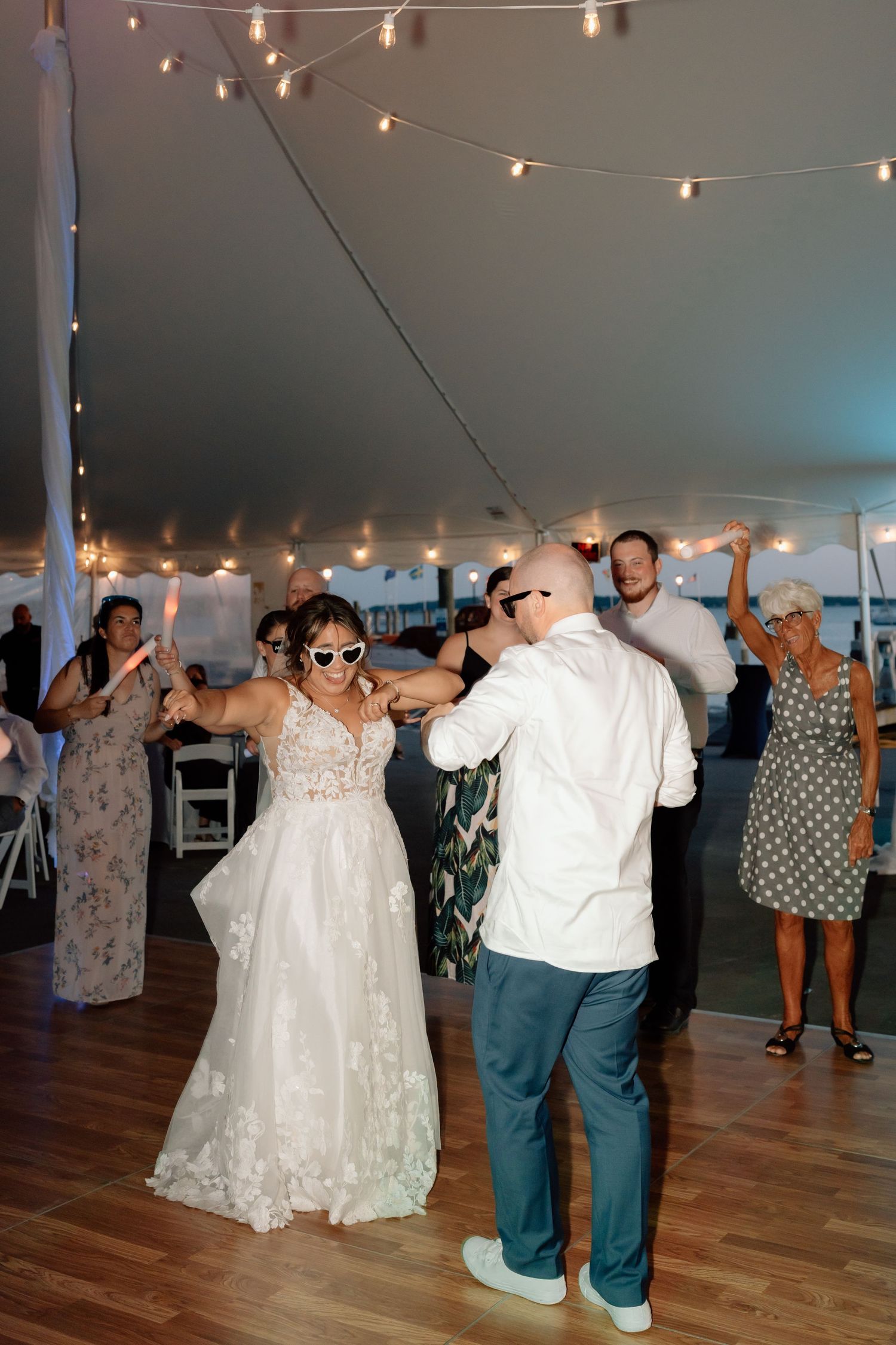 Large group dancing together at a wedding reception under a tent with string lights.