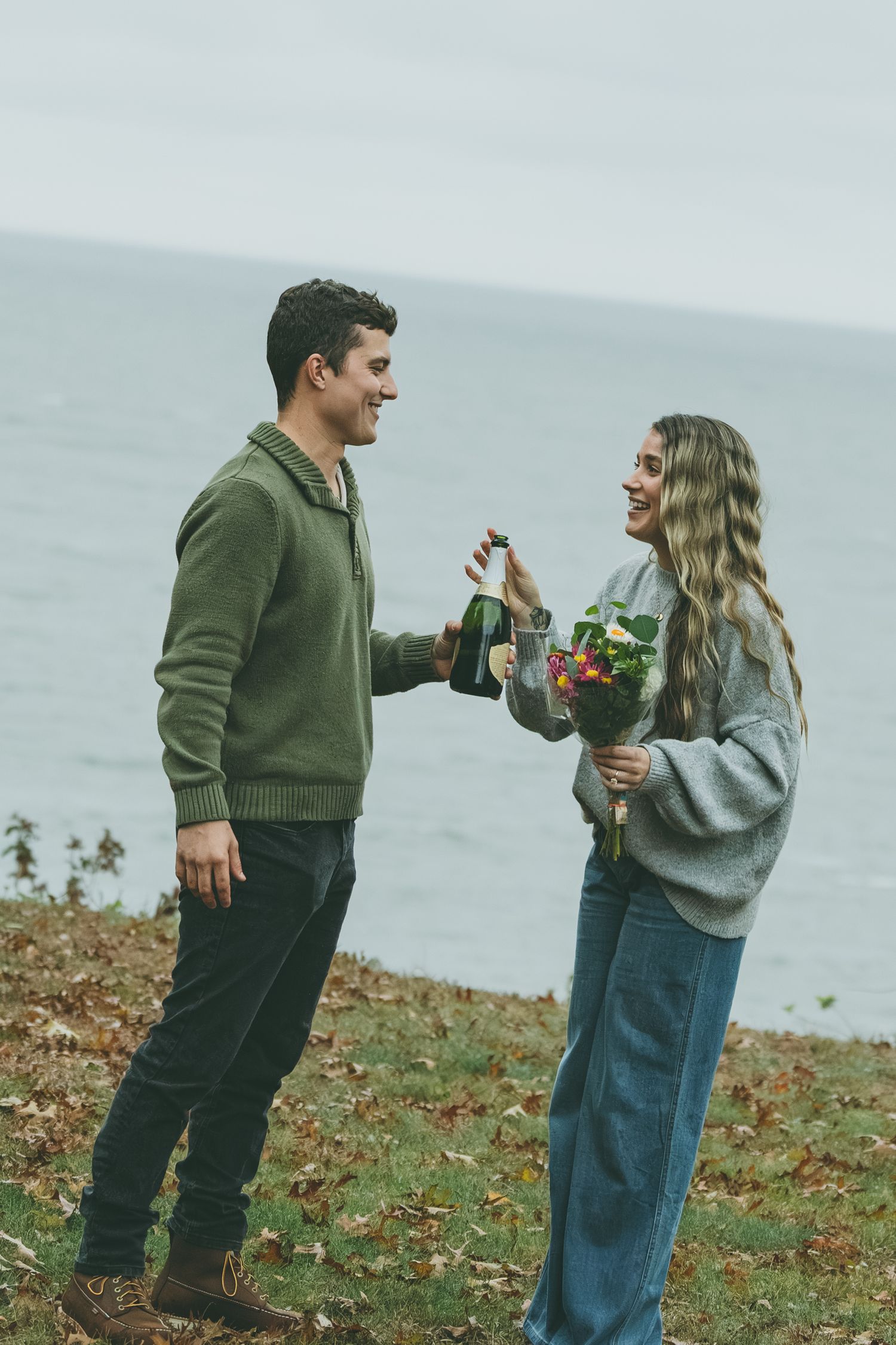 A couple shares a playful moment on a cliff overlooking the ocean during autumn.