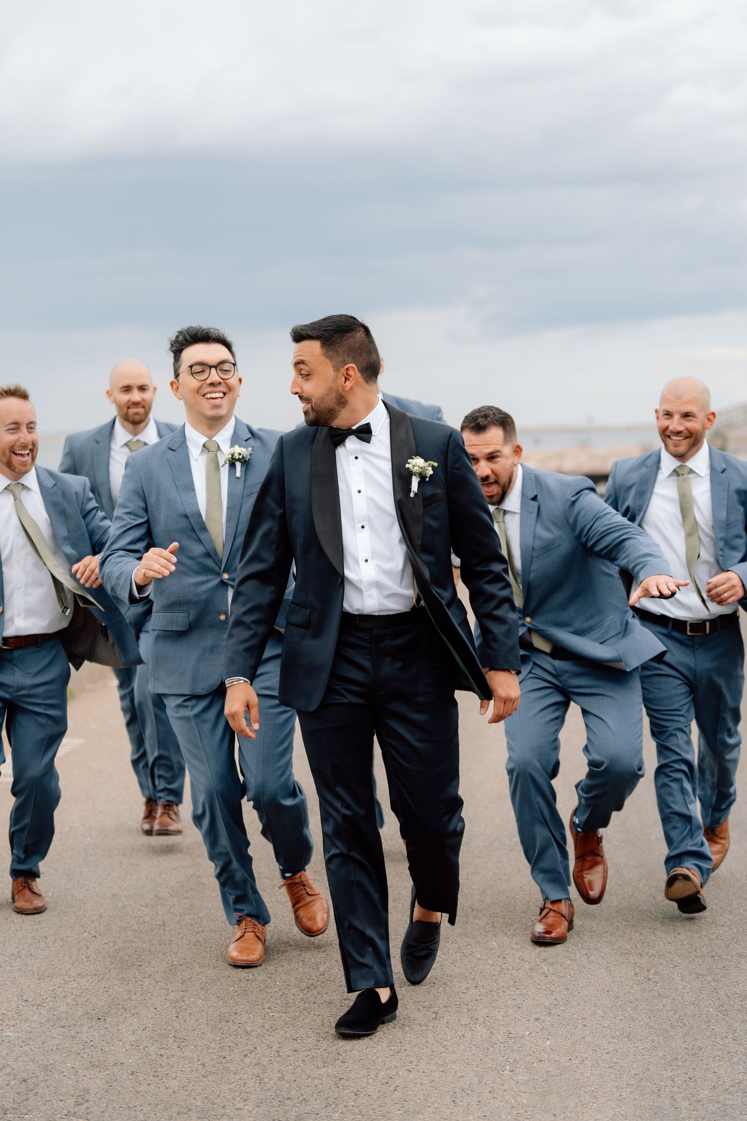 Groomsmen in blue suits walking and laughing with groom in black tuxedo.