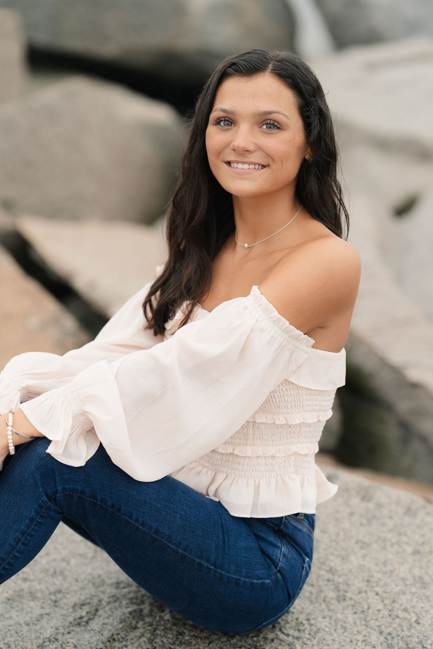 Portrait of someone in a white off-shoulder top and jeans sitting on rocks.