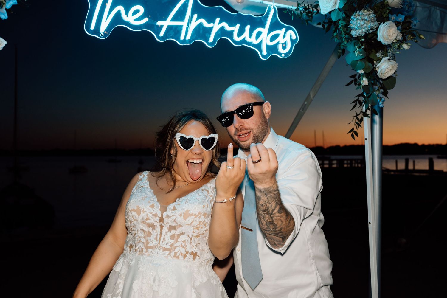 Wedding couple wearing sunglasses posing under neon sign at sunset.