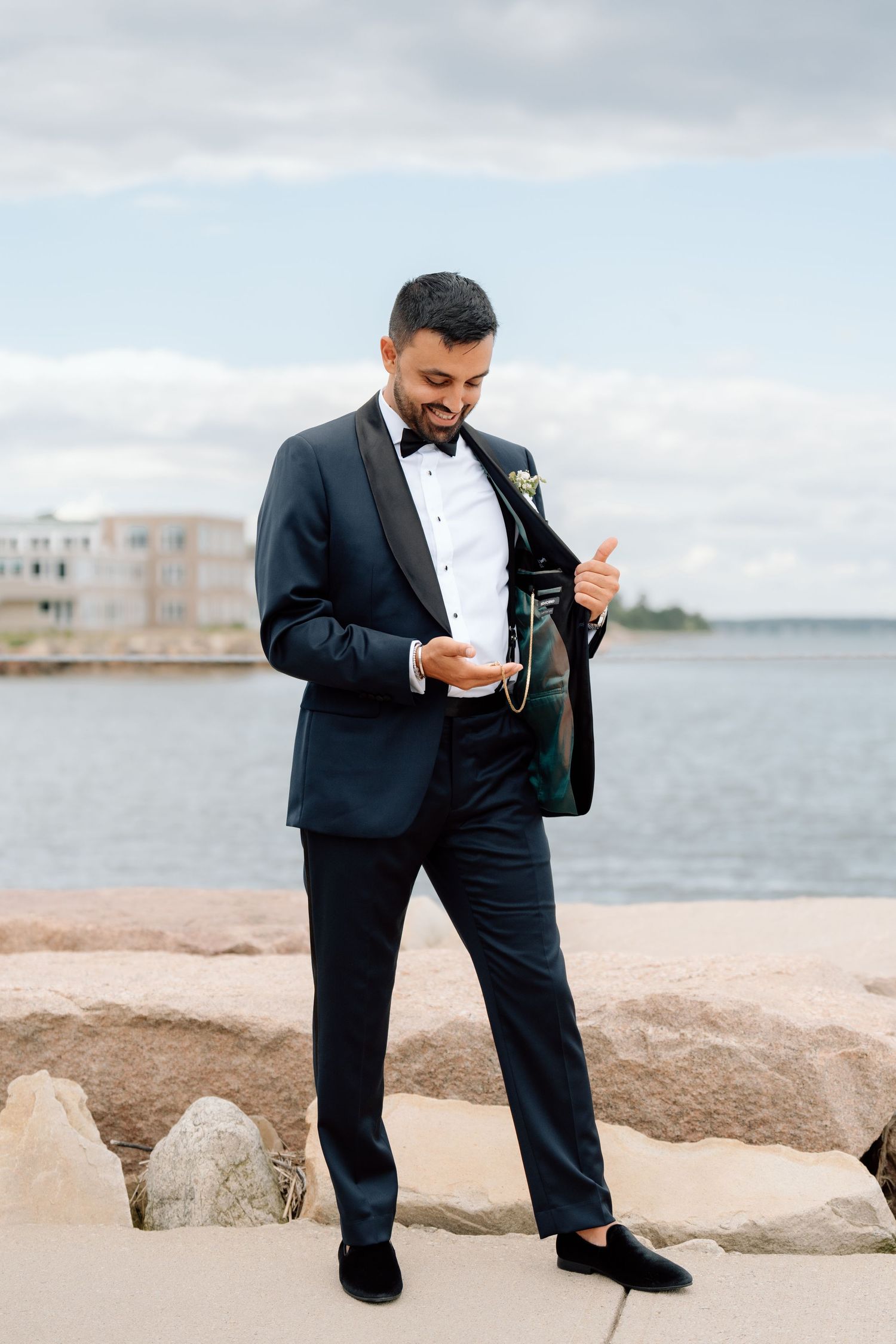 Groom in navy tuxedo standing by waterfront checking inside jacket pocket.