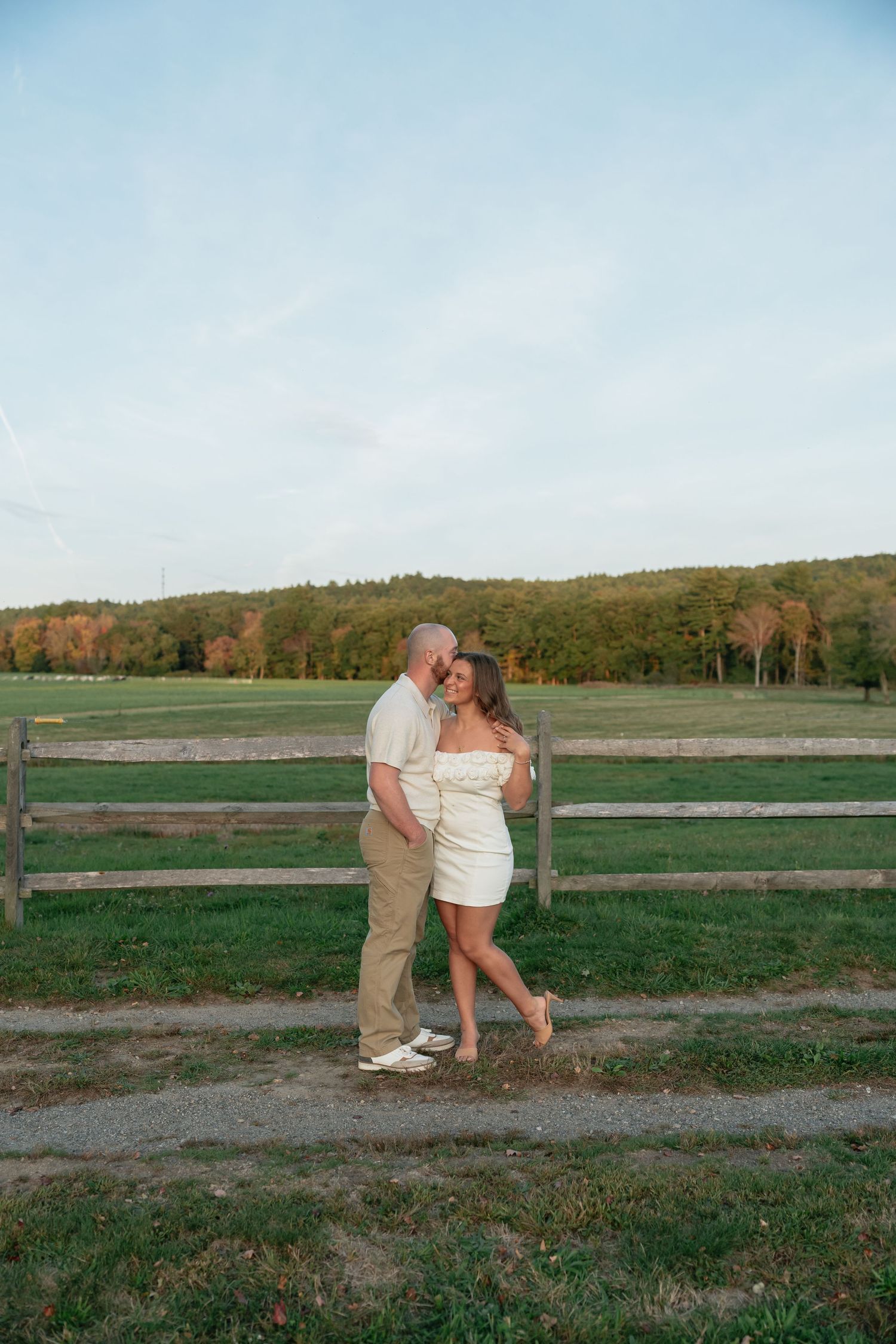 A couple shares intimate moments by a rustic wooden fence in a green countryside setting.