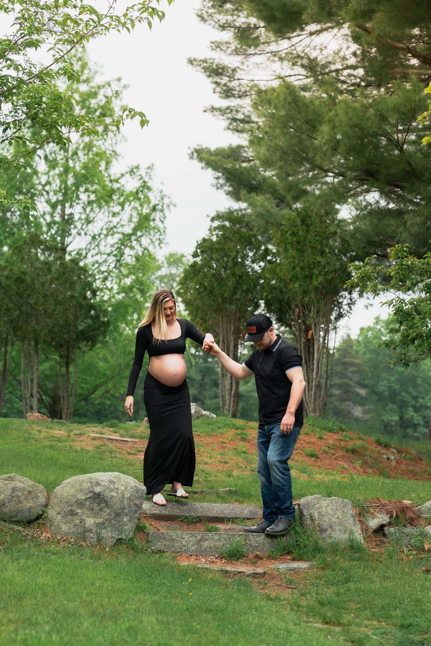 A couple poses for maternity photos on large rocks surrounded by pine trees and green grass.