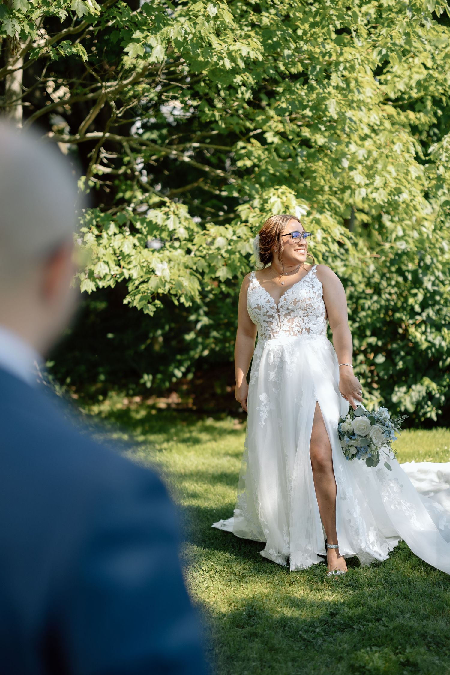 A bride in a flowing white gown holds a bouquet in a sunny garden setting.