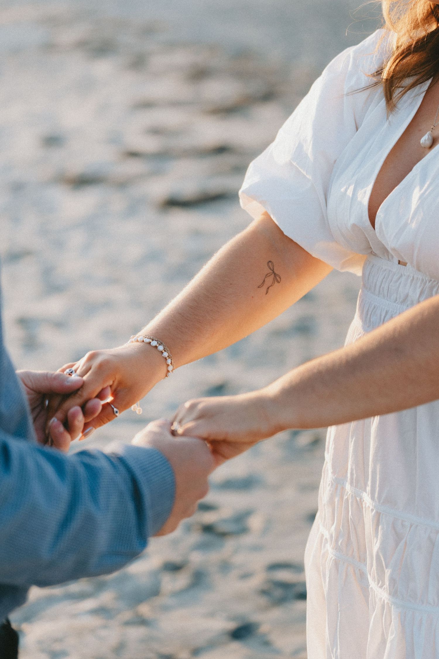 Two people hold hands tenderly on a sandy beach at sunset.