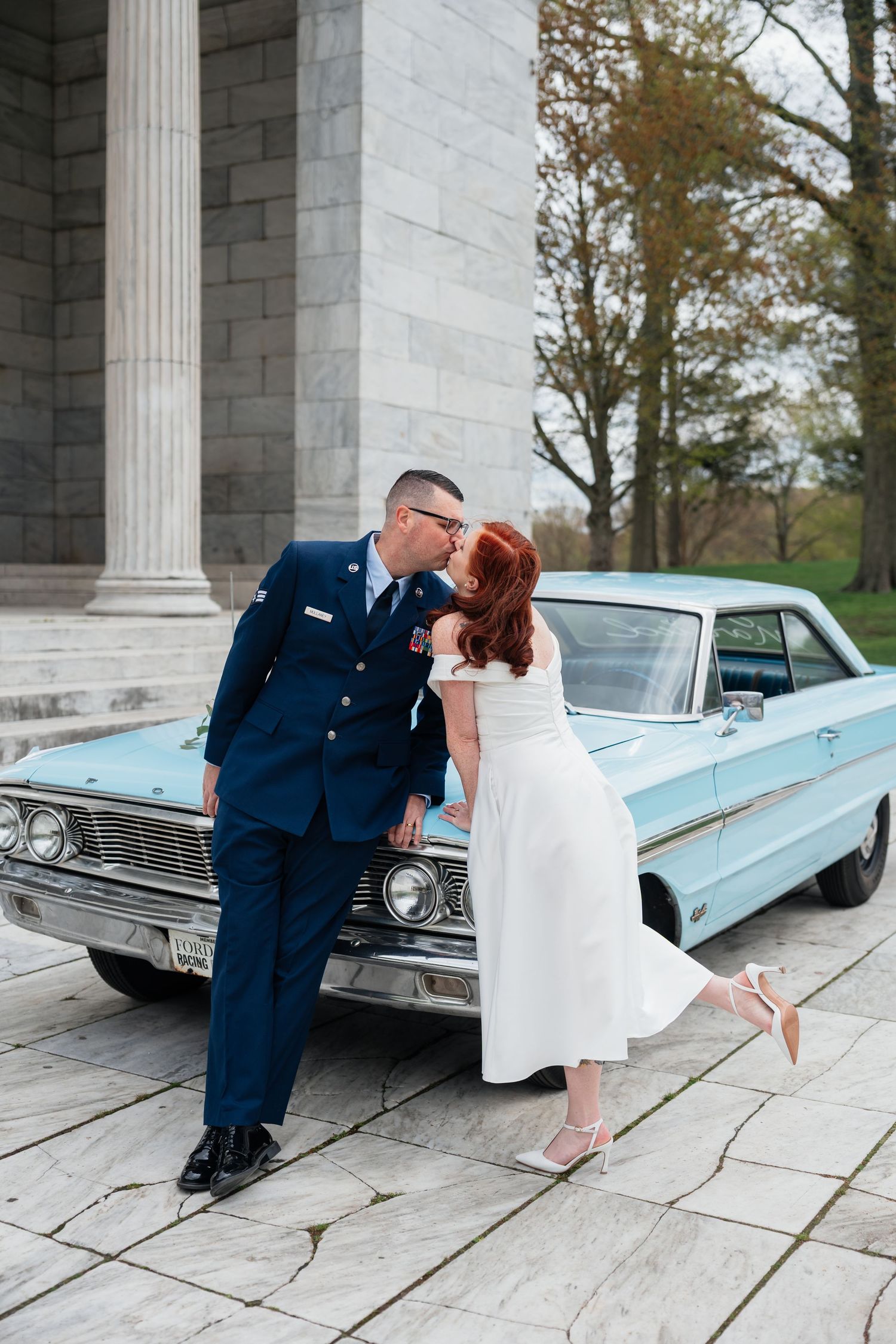 Newlyweds sharing intimate moment beside classic blue car at stone pavilion.