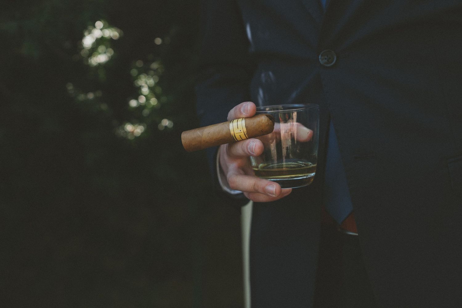 A hand holding a glass of whiskey and cigar against a dark background with bokeh lights.