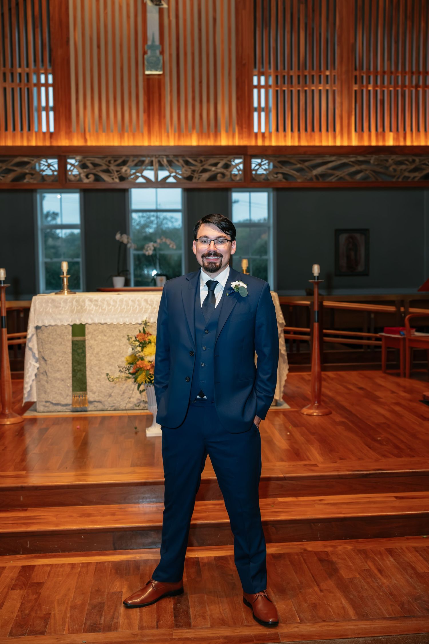 Groom poses in navy suit before altar in wooden church interior.