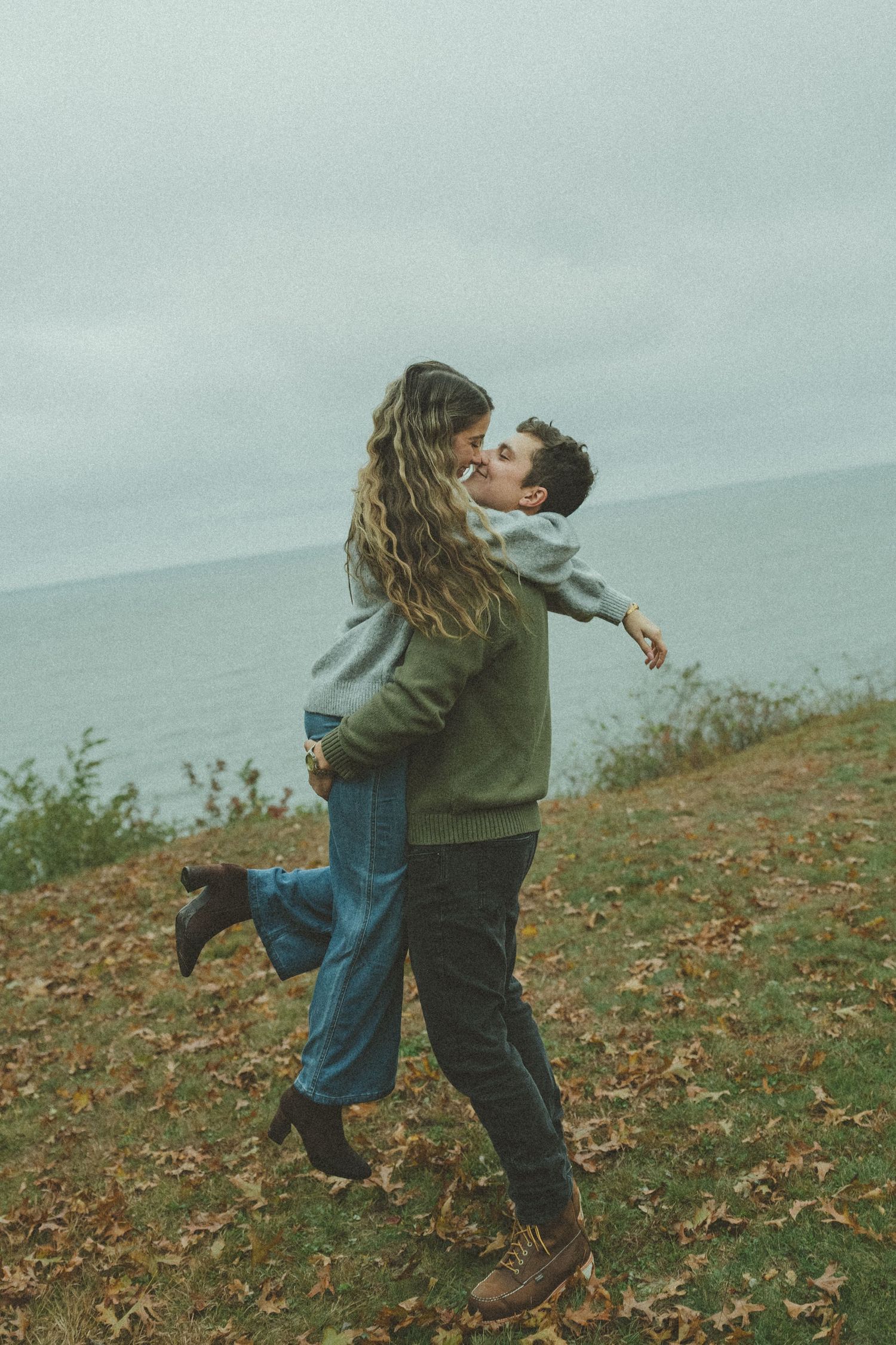 A couple playfully spins together on a hillside covered in autumn leaves.