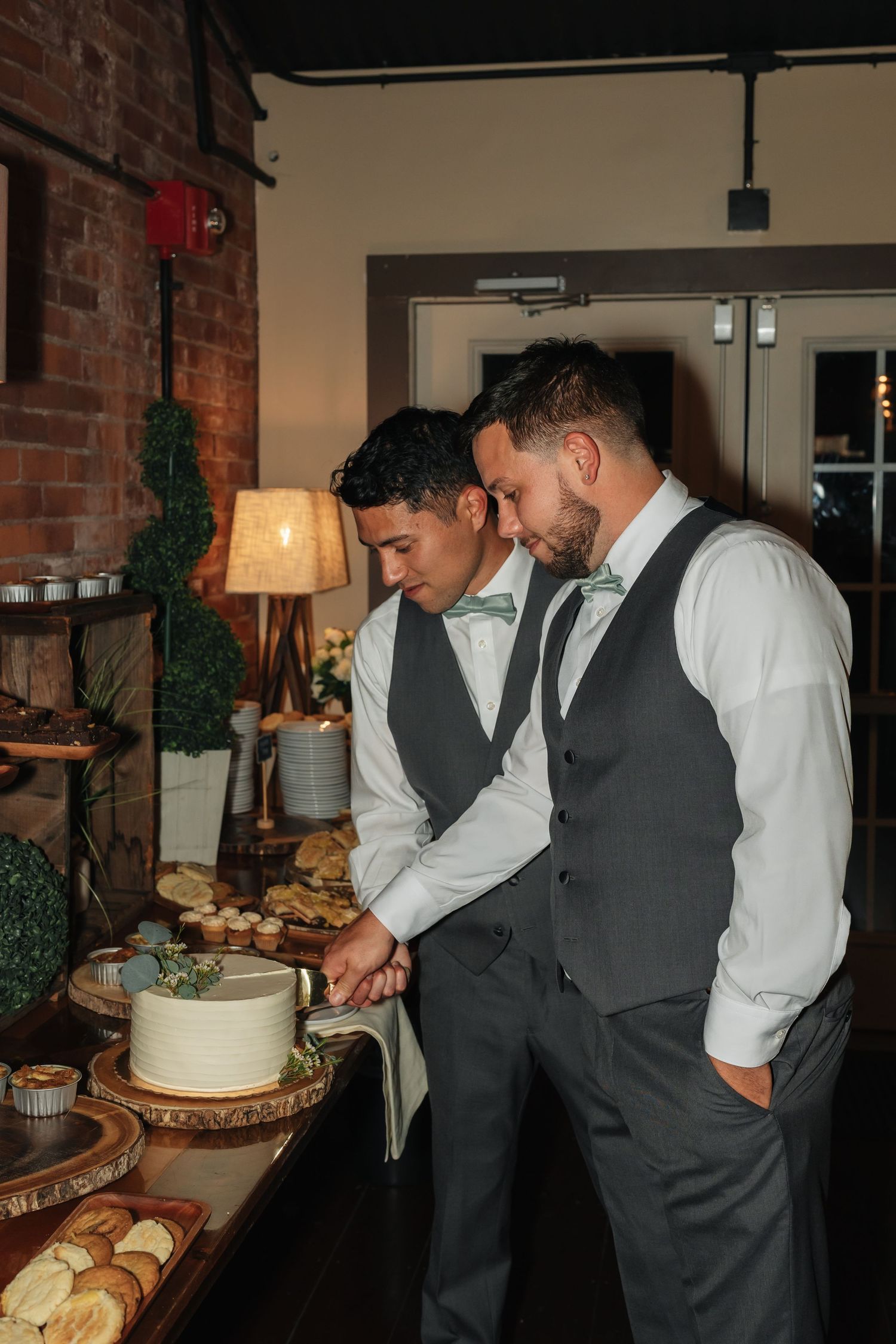 Groomsmen in grey vests examining dessert display at wedding reception buffet table.
