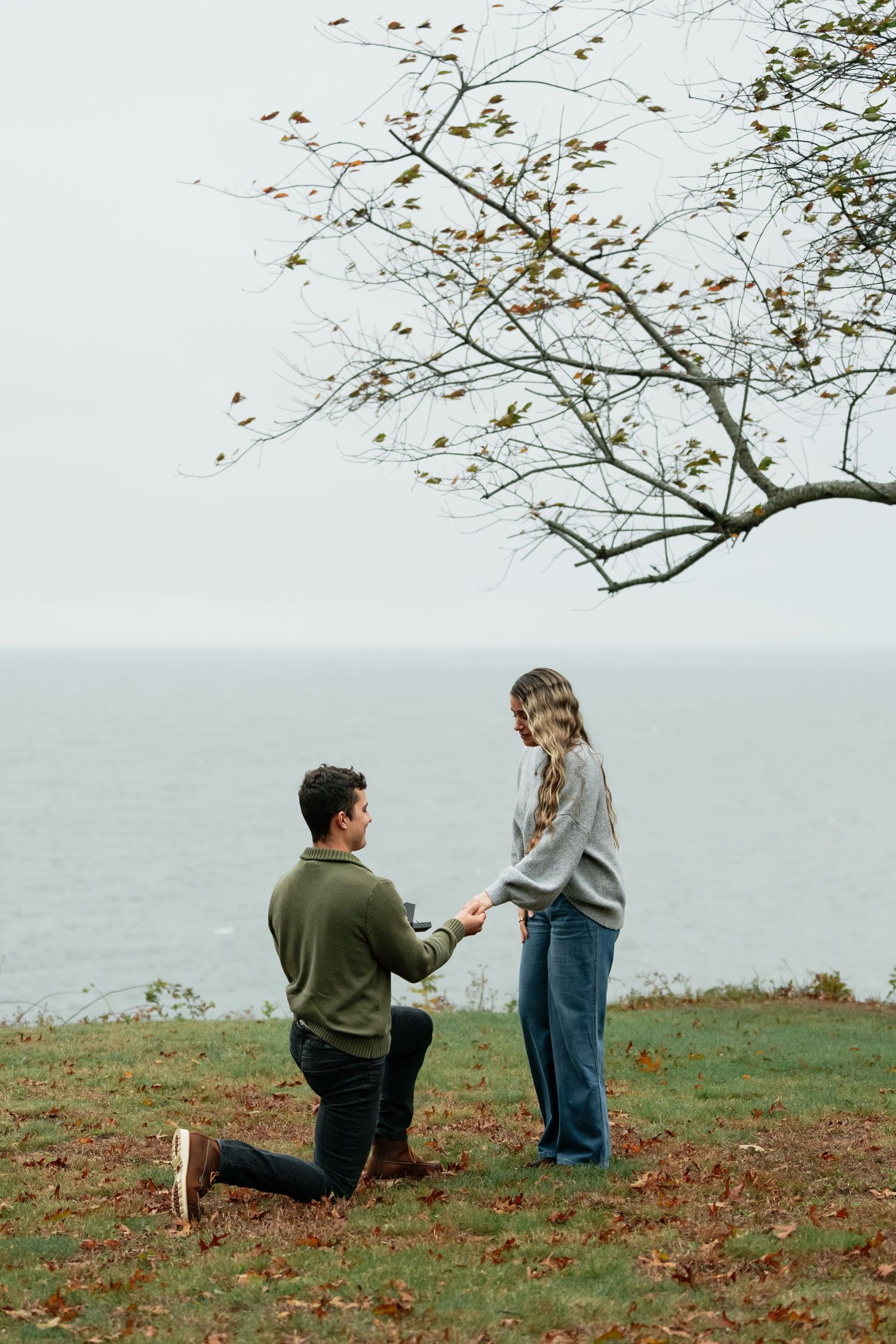 Scenic autumn proposal scene with couple by water under bare trees.