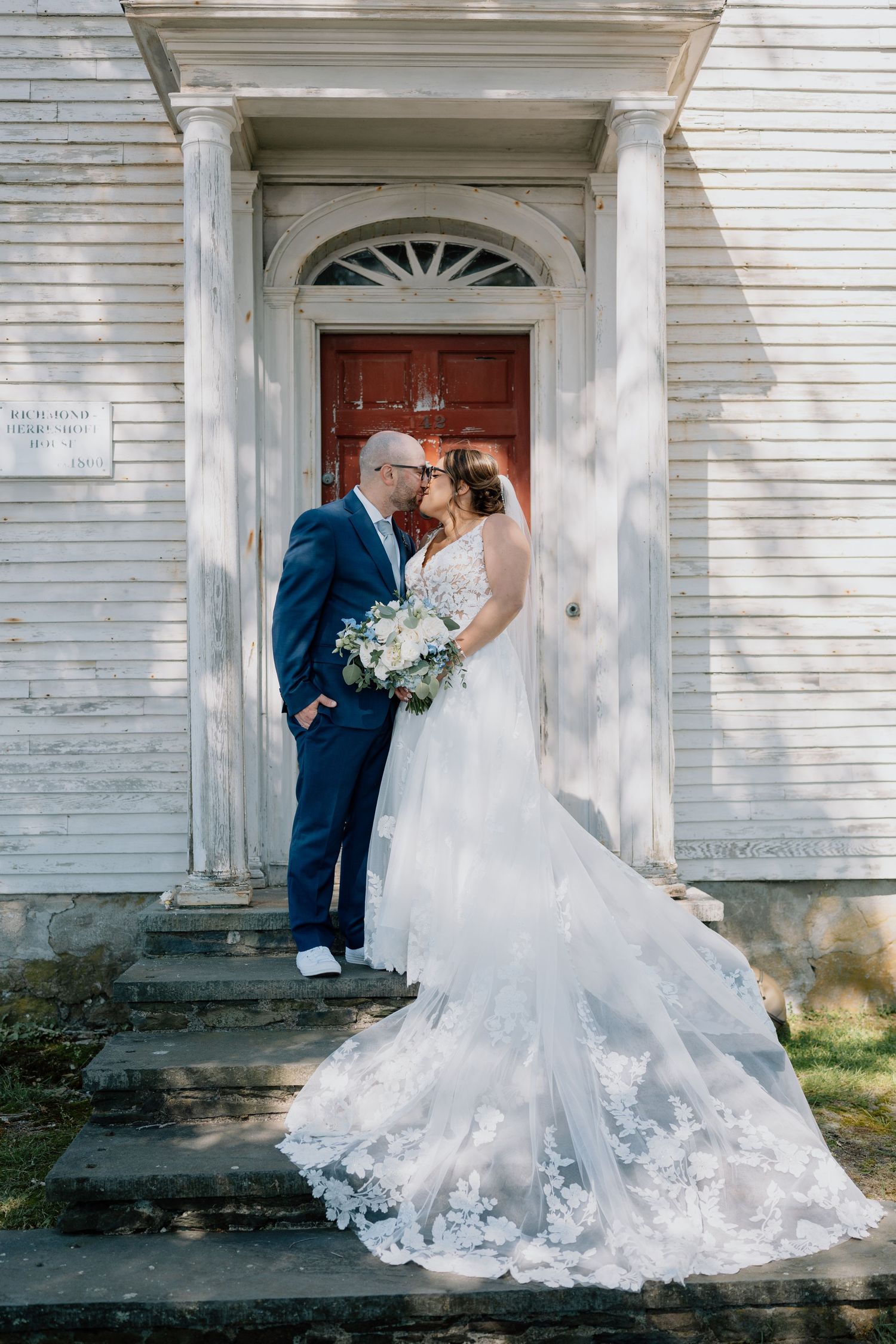 A wedding couple poses in front of a classic white church doorway with ornate architectural details.