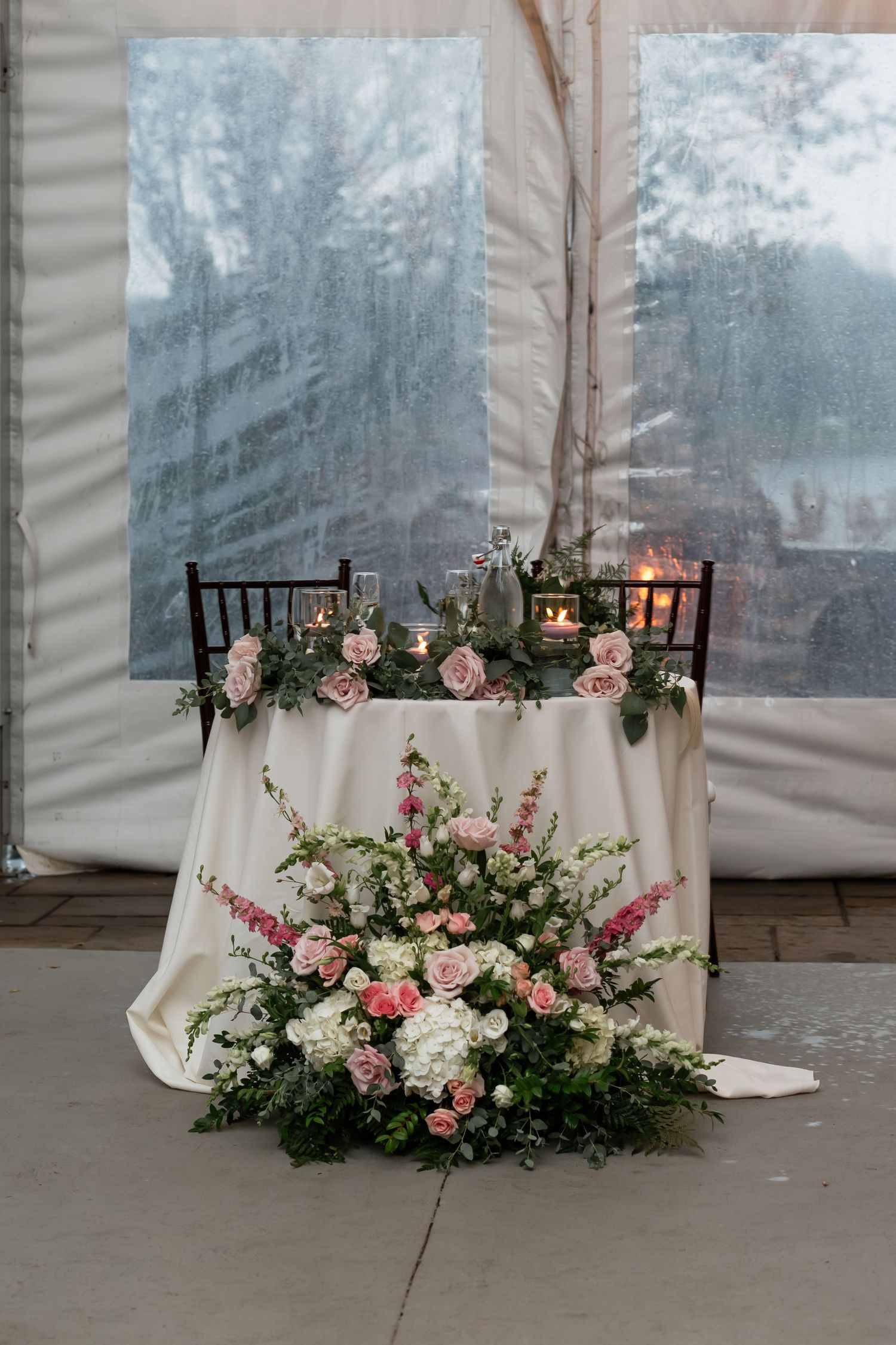 Elegant sweetheart table decorated with pink roses and greenery against white tent backdrop.