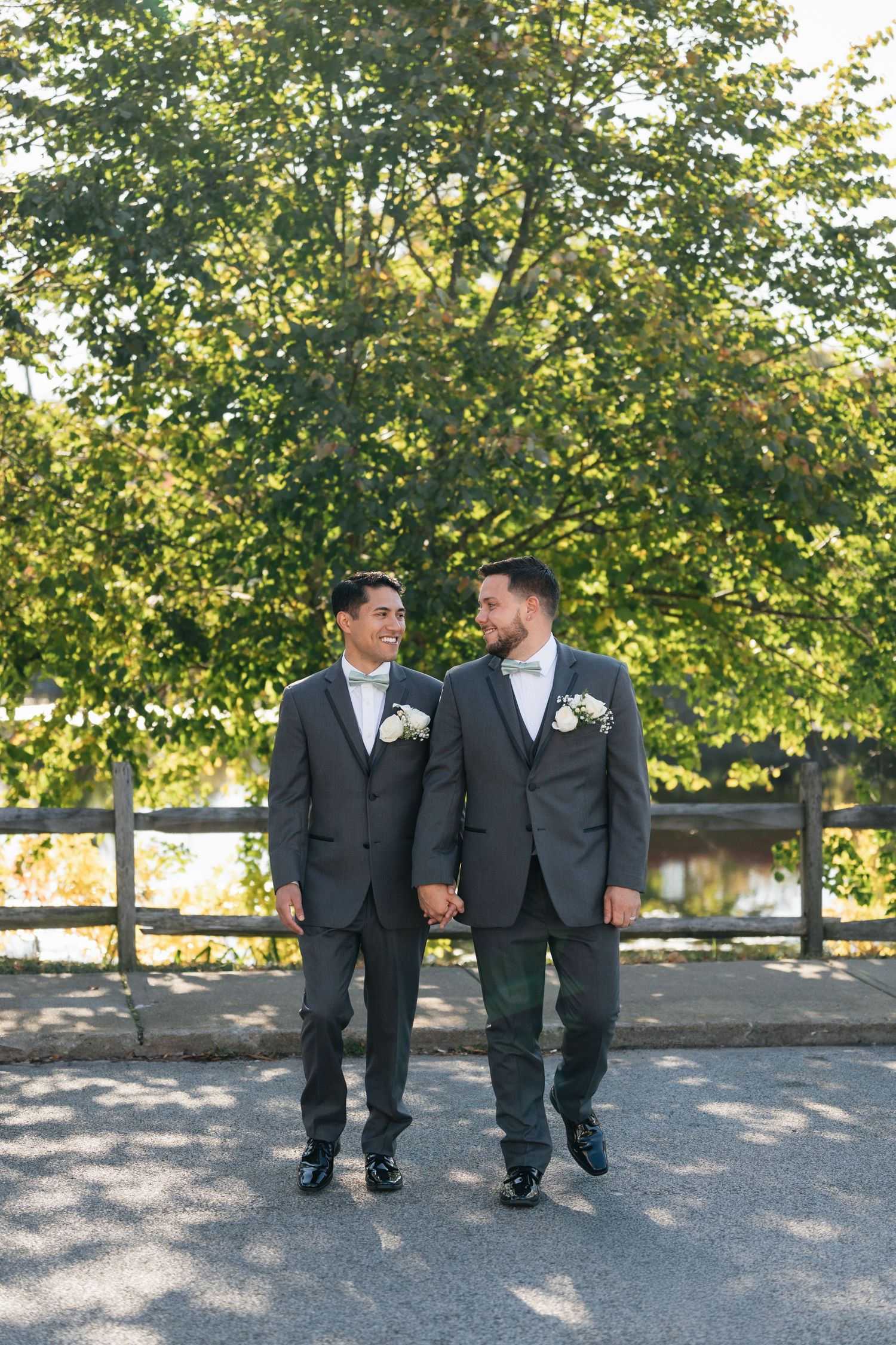Newlyweds in coordinating gray suits walk hand in hand along a tree-lined path.