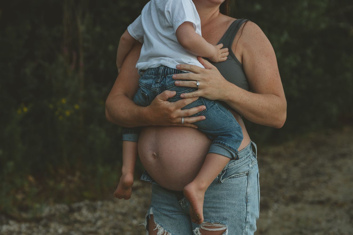 A tender maternity moment showing a pregnant woman holding a toddler against natural backdrop.