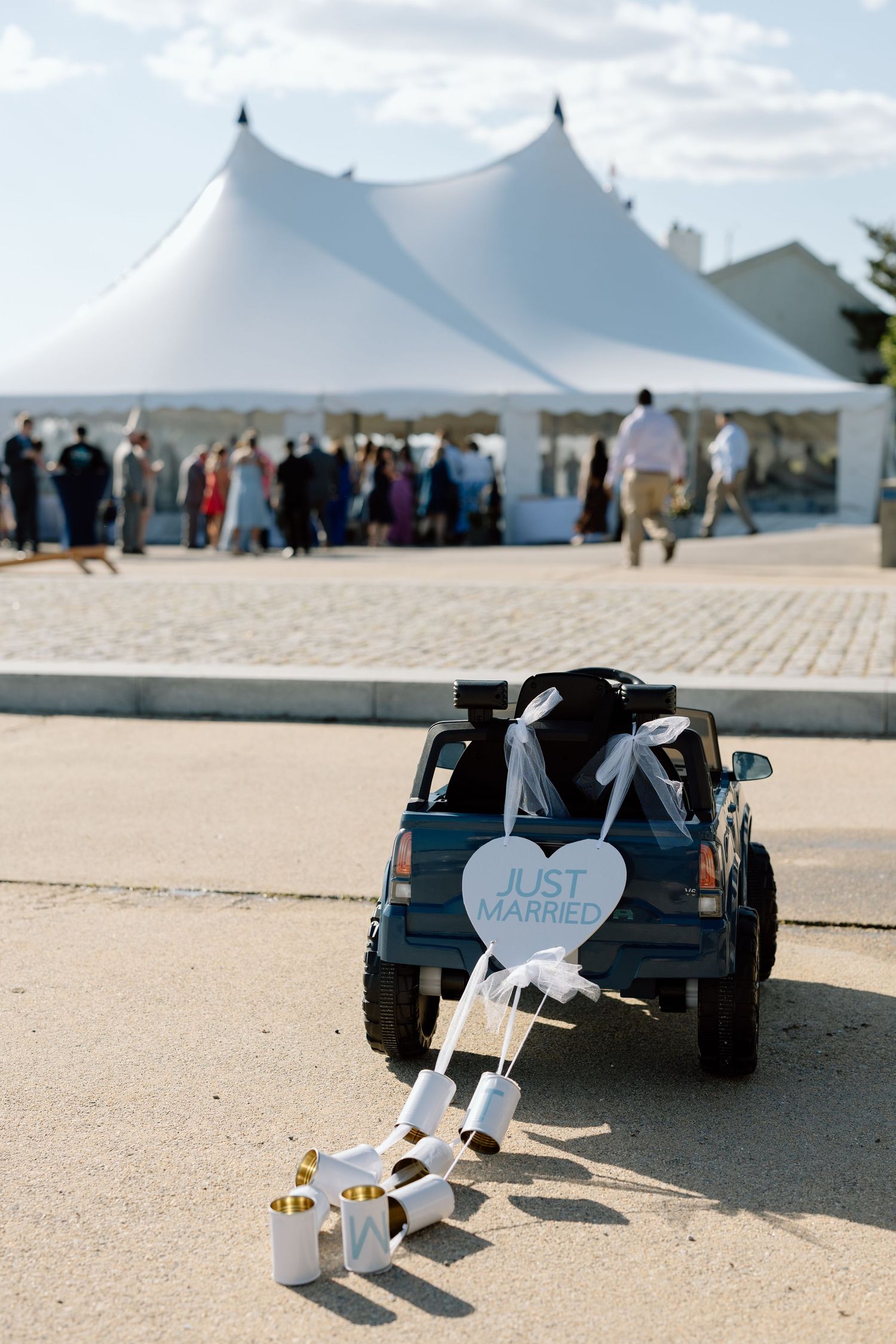Toy cars with 'Just Married' decorations on sandy beach with white tents in background.