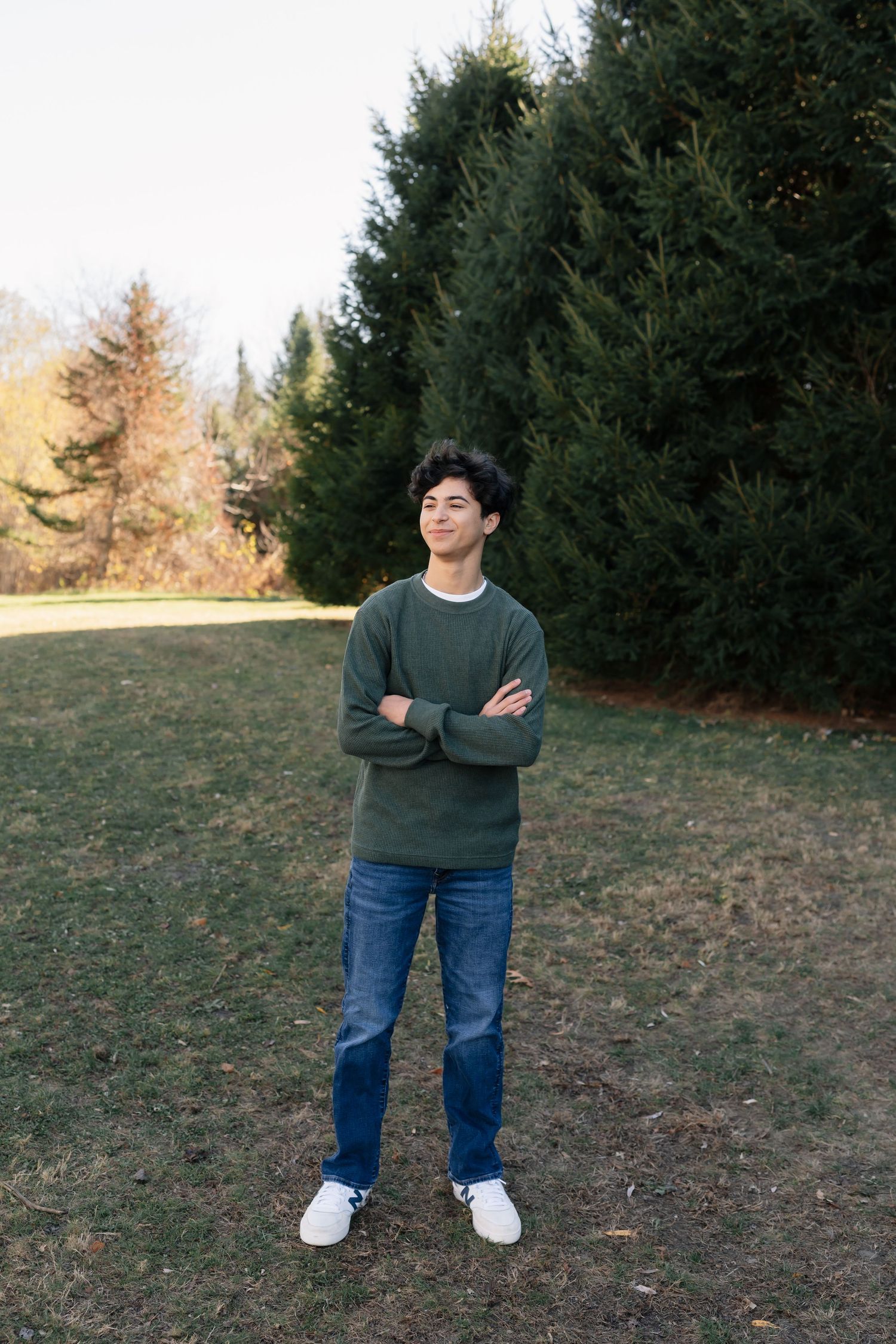 Person in dark sweater and jeans posing casually near evergreen trees.