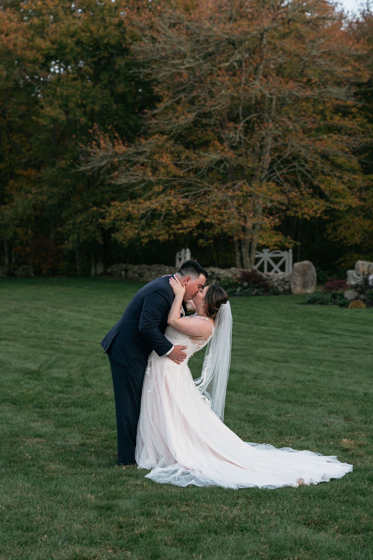 Romantic wedding couple pose in grassy field with autumn trees.