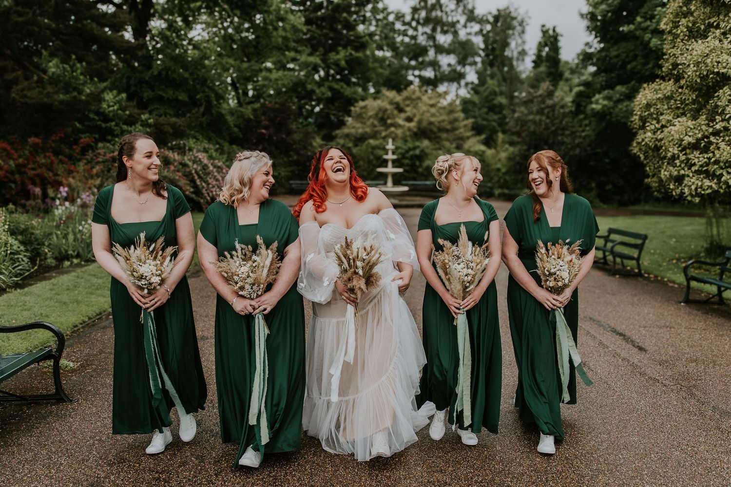 Bride in white gown poses with bridesmaids wearing matching emerald green dresses in garden setting.