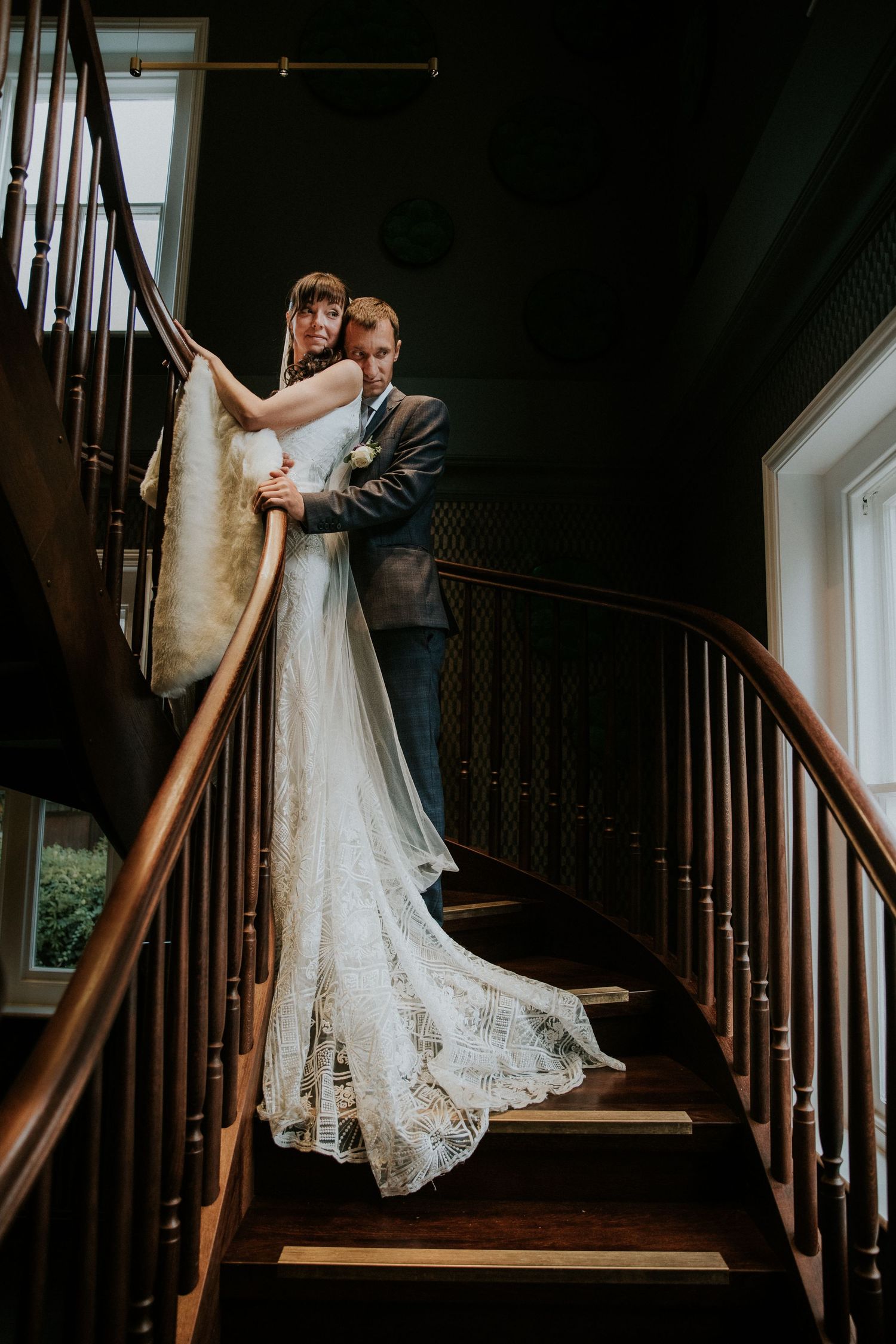 A romantic wedding couple pose on an elegant wooden staircase with intricate lace dress details.