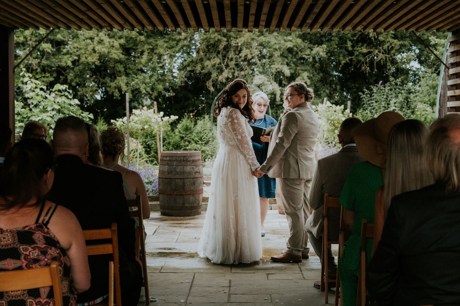 Outdoor wedding ceremony under wooden pergola with rustic wine barrel decor.