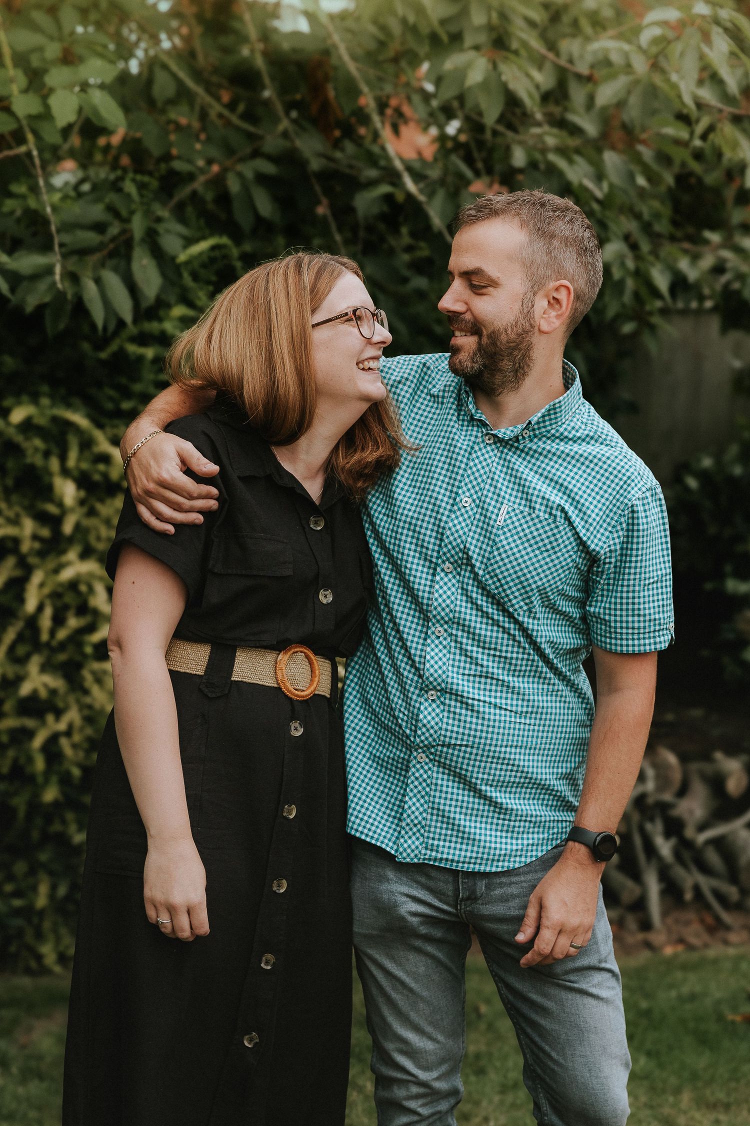 A couple shares a tender moment in a garden while wearing a black dress and teal checkered shirt.