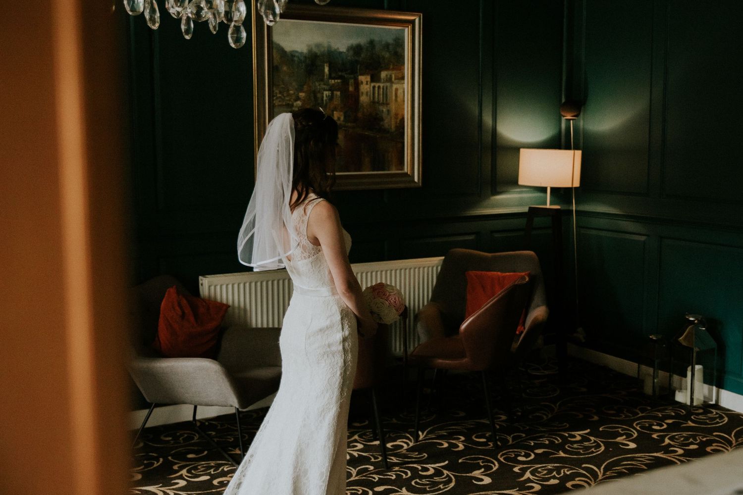 A bride in a fitted white wedding gown stands elegantly in a dark green room with vintage decor.