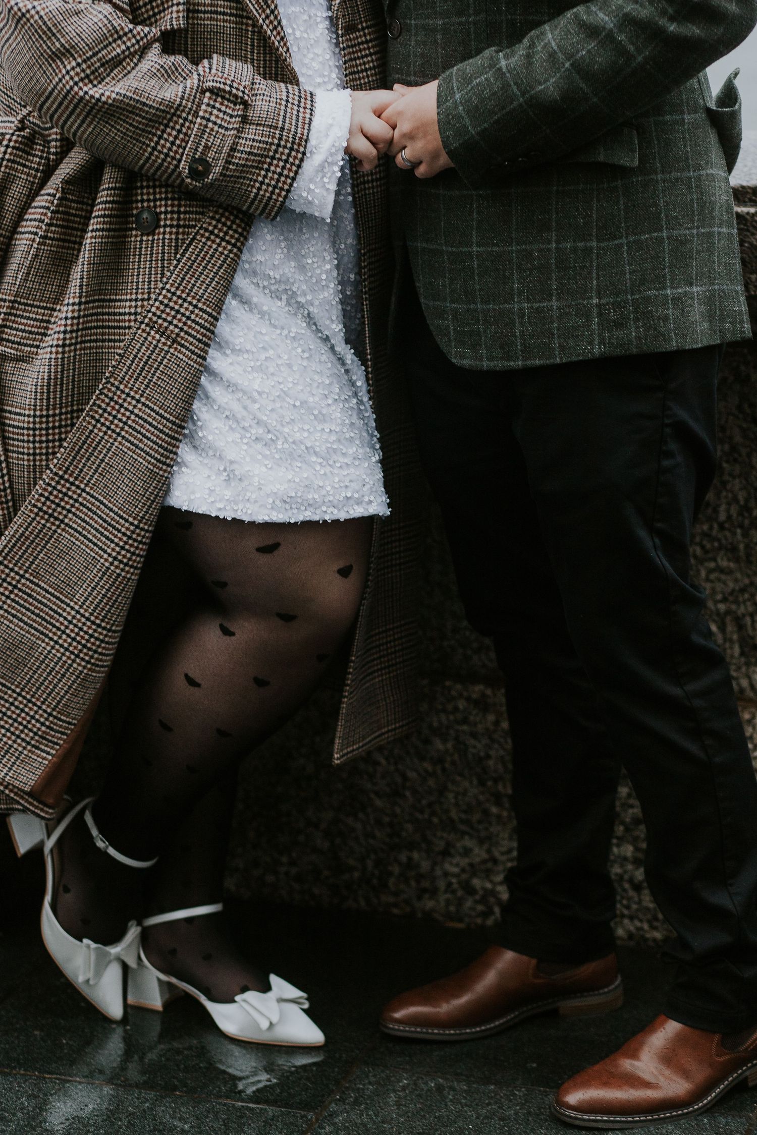 Close-up of two people's feet and hands touching, showing patterned coat and dress details.