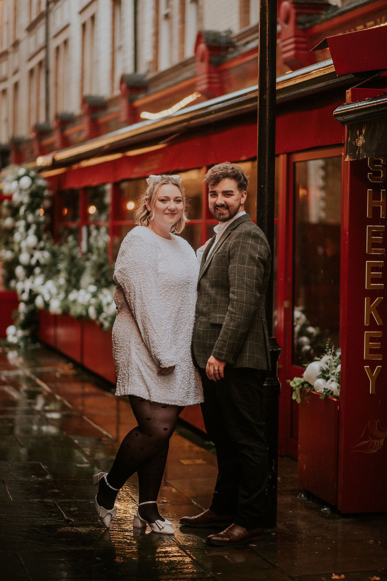 A couple poses together outside a red storefront on a rainy day in the city.