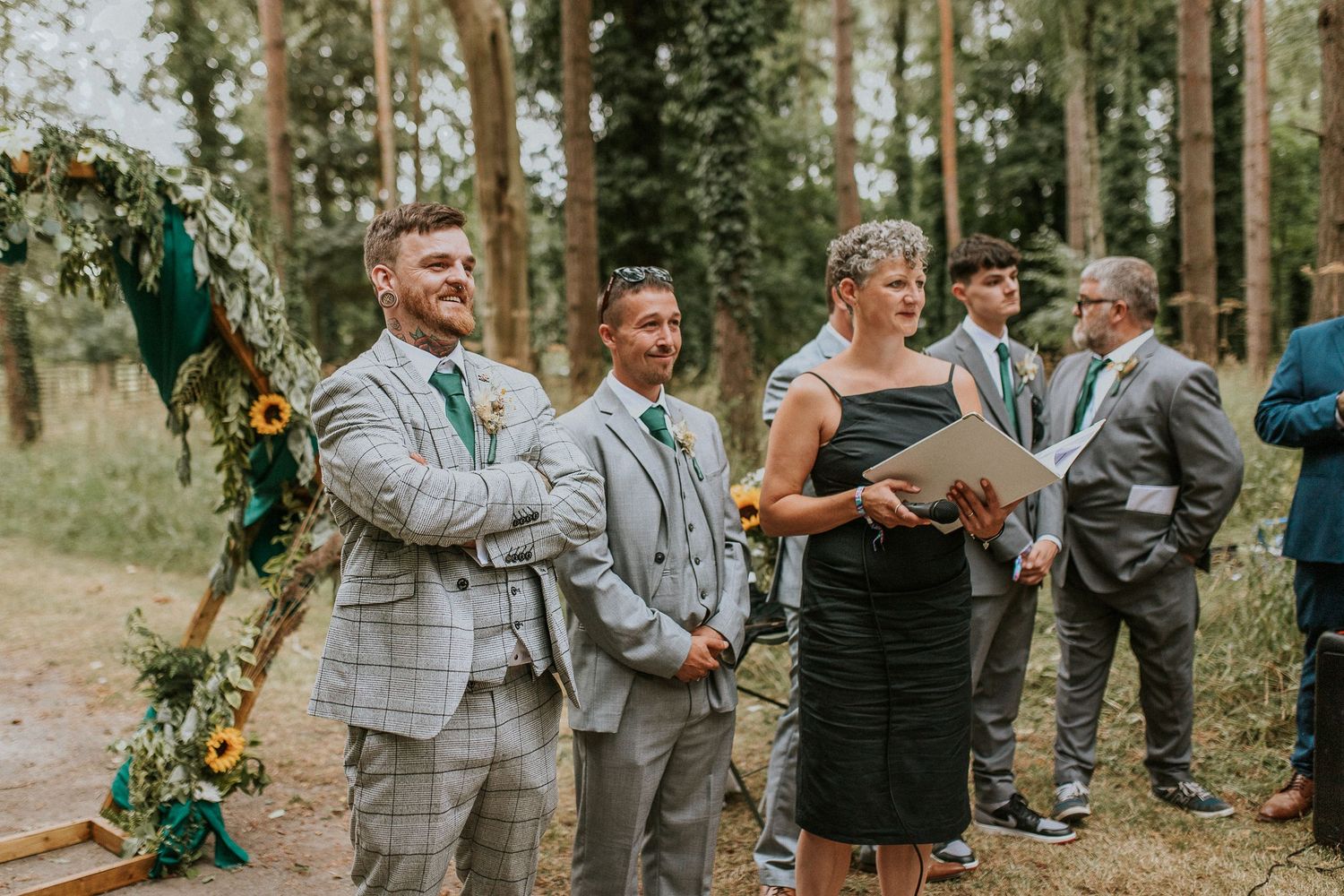 Wedding ceremony in progress with officiant and wedding party in grey suits among forest trees.