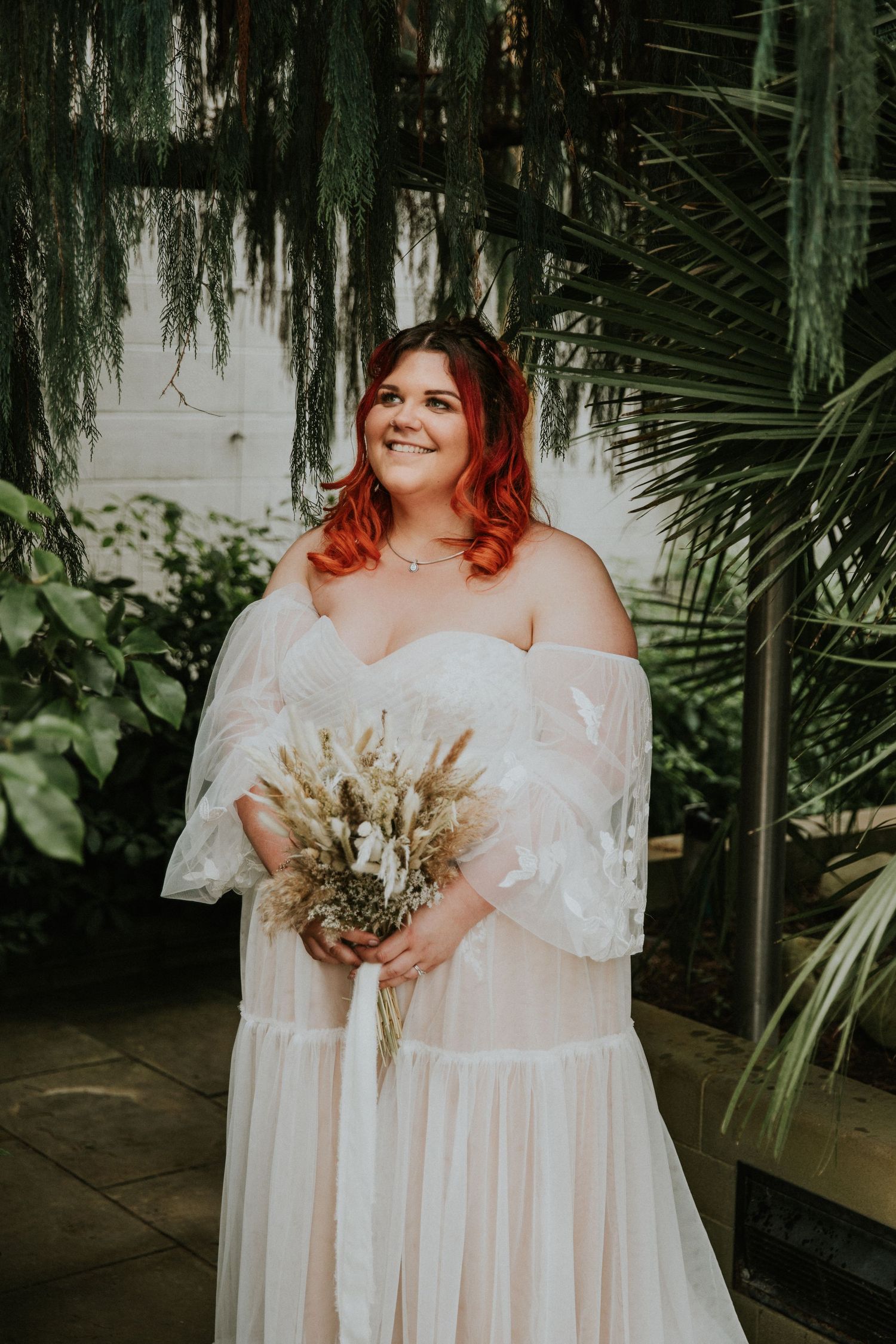 A bride in an off-shoulder wedding dress with vibrant red hair holds a dried flower bouquet in a greenhouse setting.