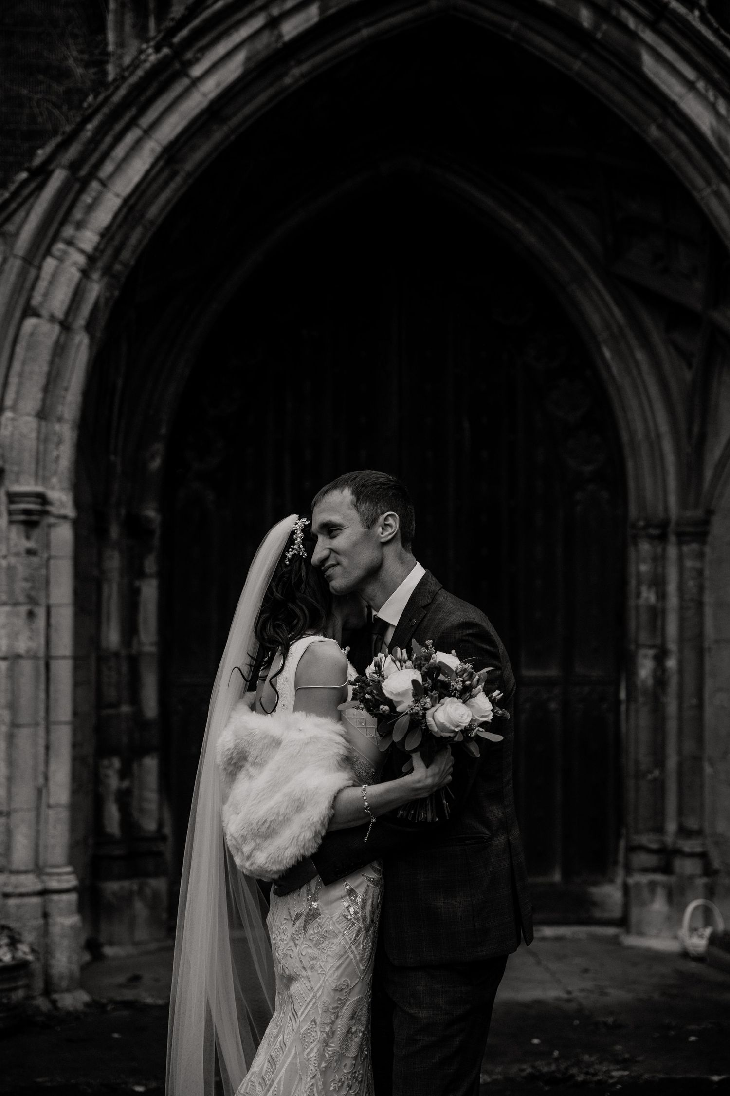 Romantic black and white portrait of newlyweds embracing under stone church archway.