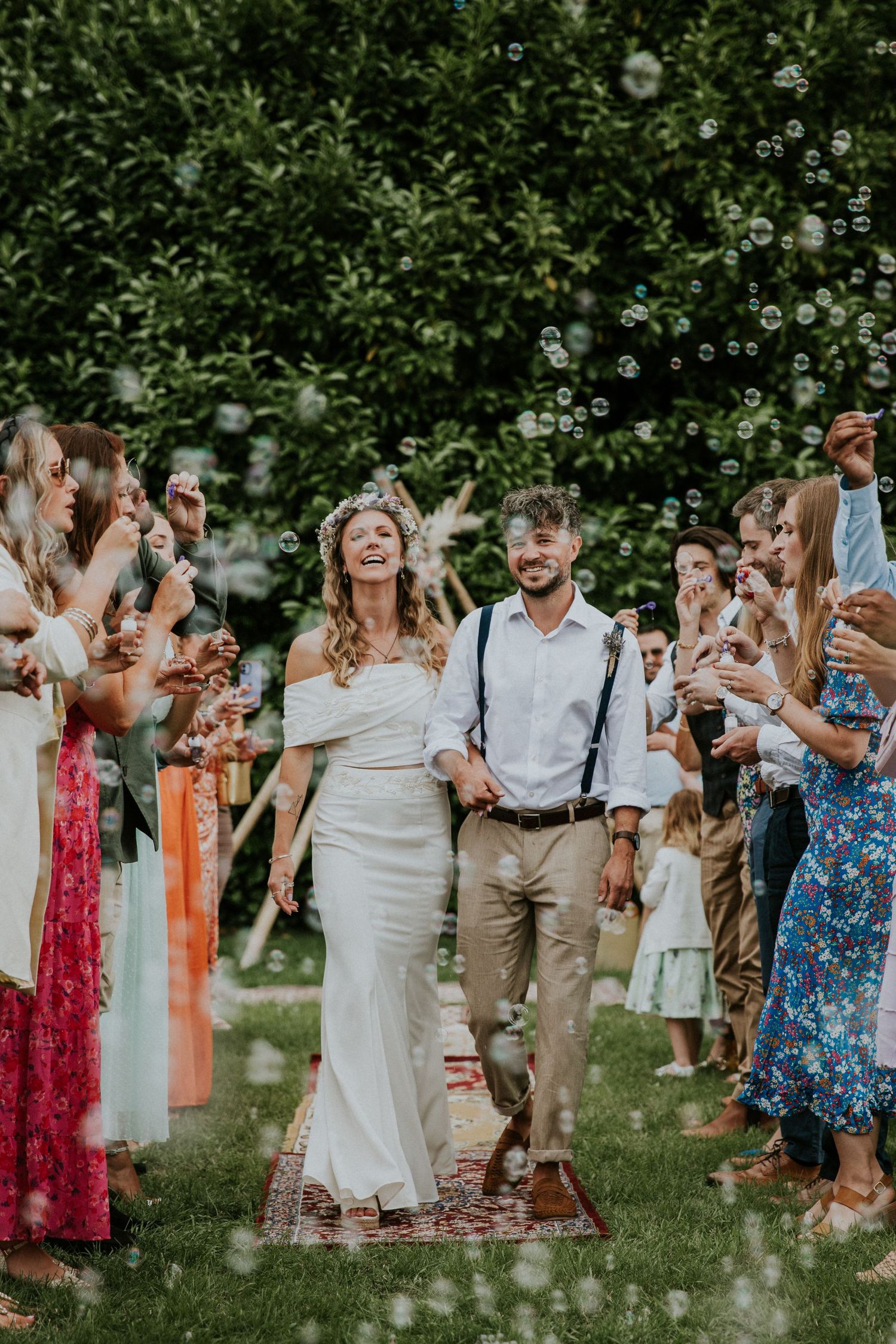 Newlyweds walk through bubble send-off surrounded by colorfully dressed guests in a garden setting.