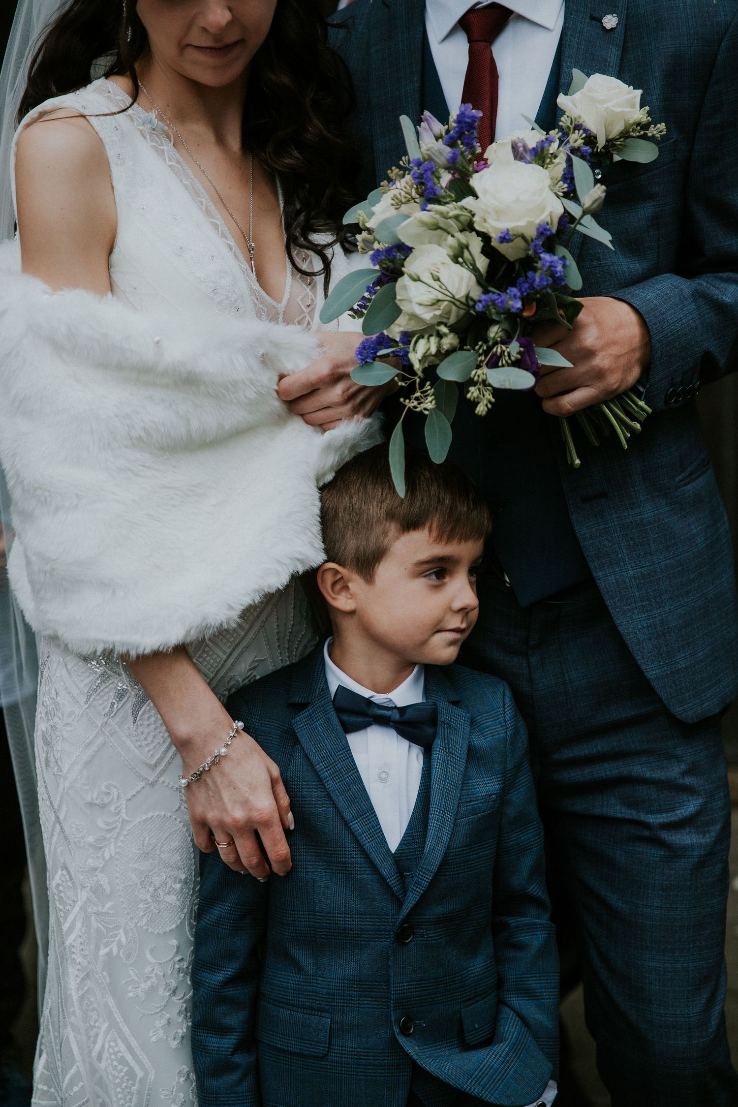 Young ring bearer in navy suit stands between bride and groom holding elegant floral bouquet.