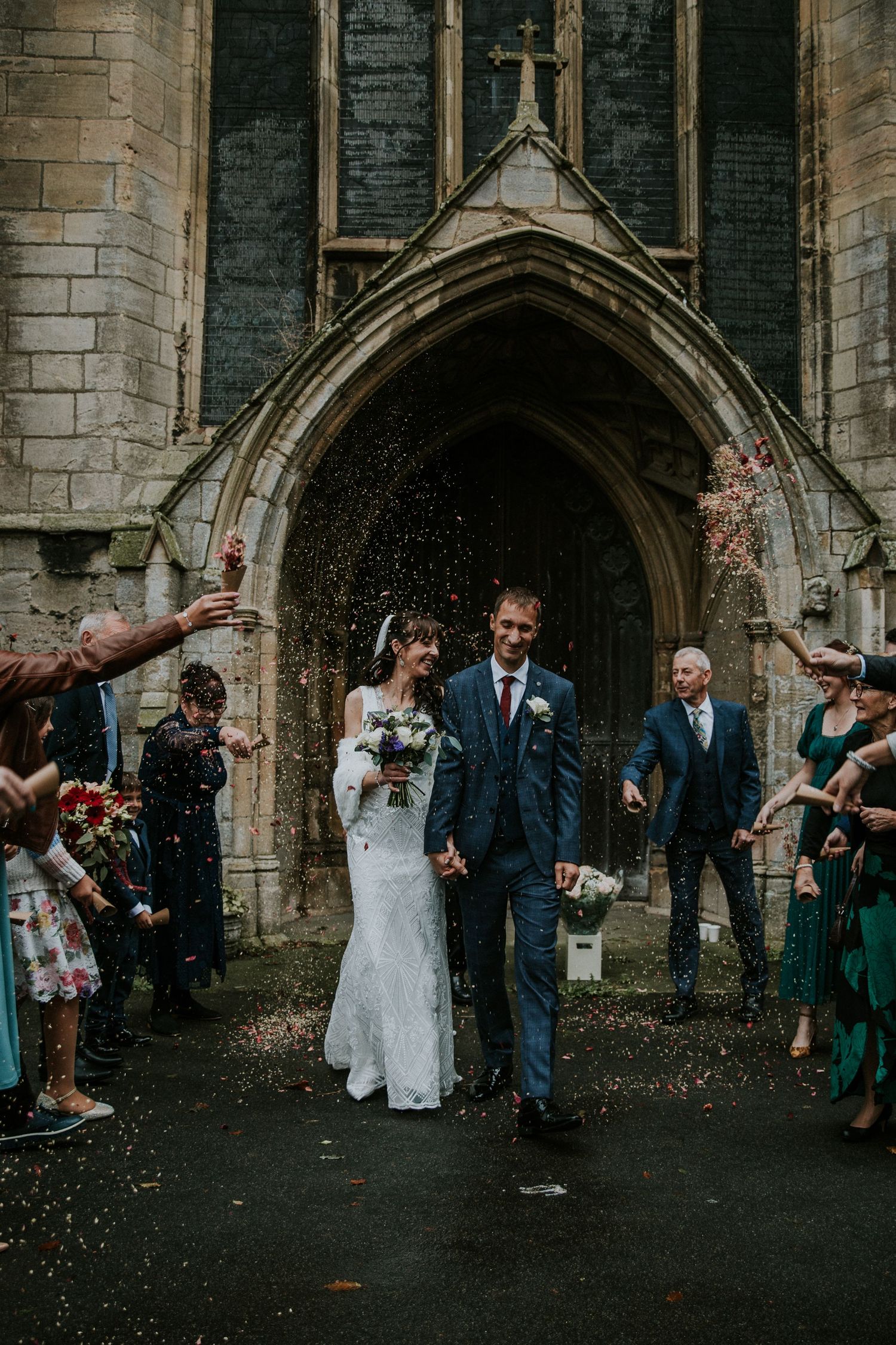 Newlyweds exit gothic church archway while guests throw confetti in celebration.