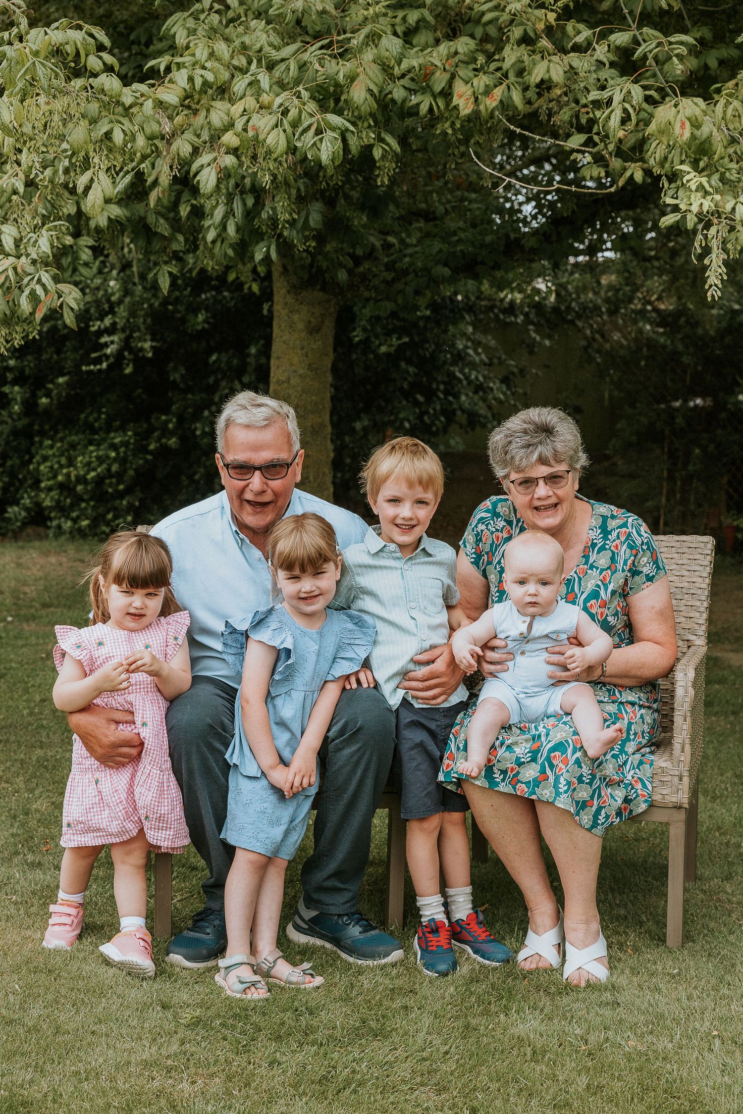 Family group sits together on a wooden bench in a lush garden setting.
