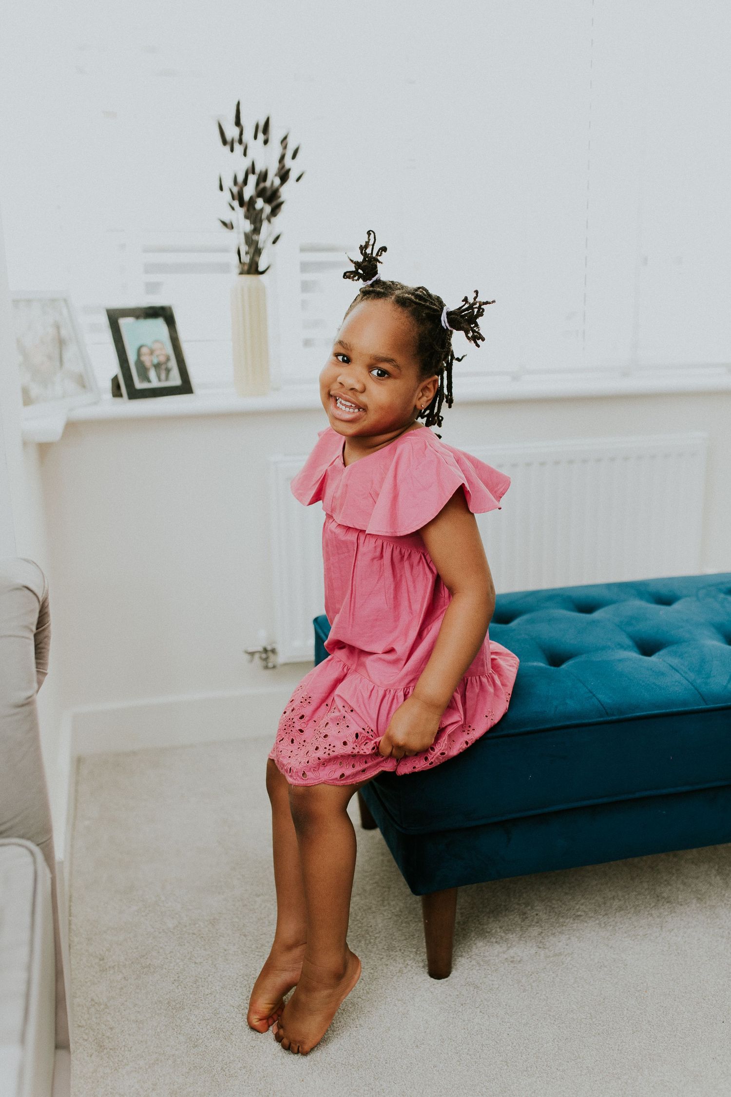 Young child wearing a pink ruffled dress plays on a blue tufted bench in a bright white room.