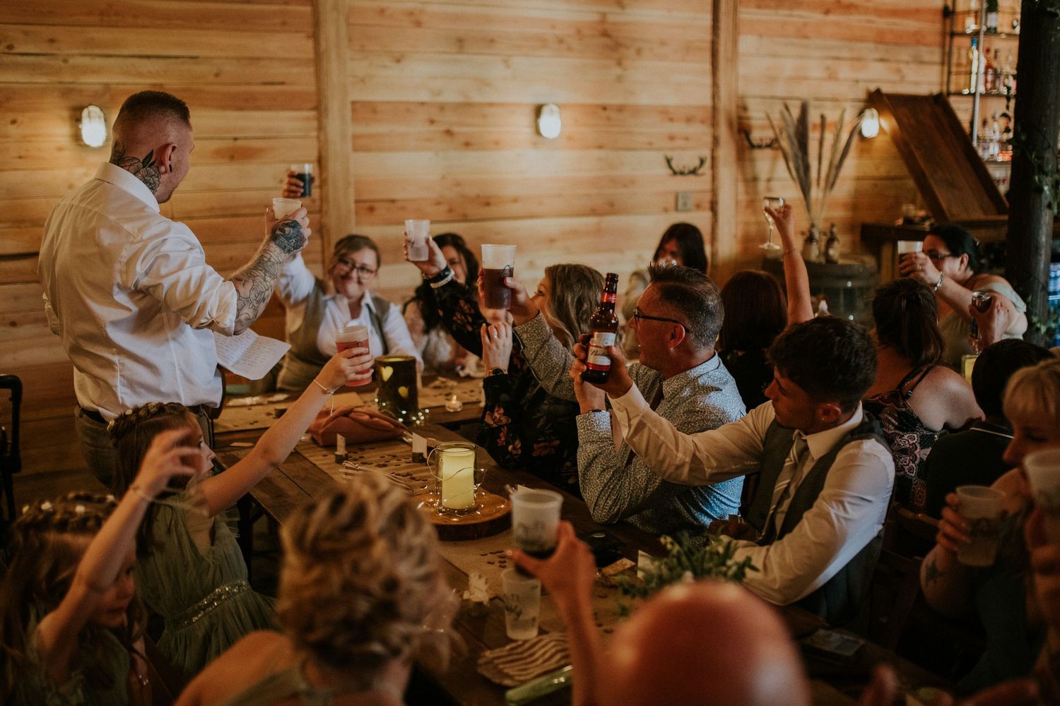 Wedding guests raise their glasses during a toast in a rustic wooden venue.