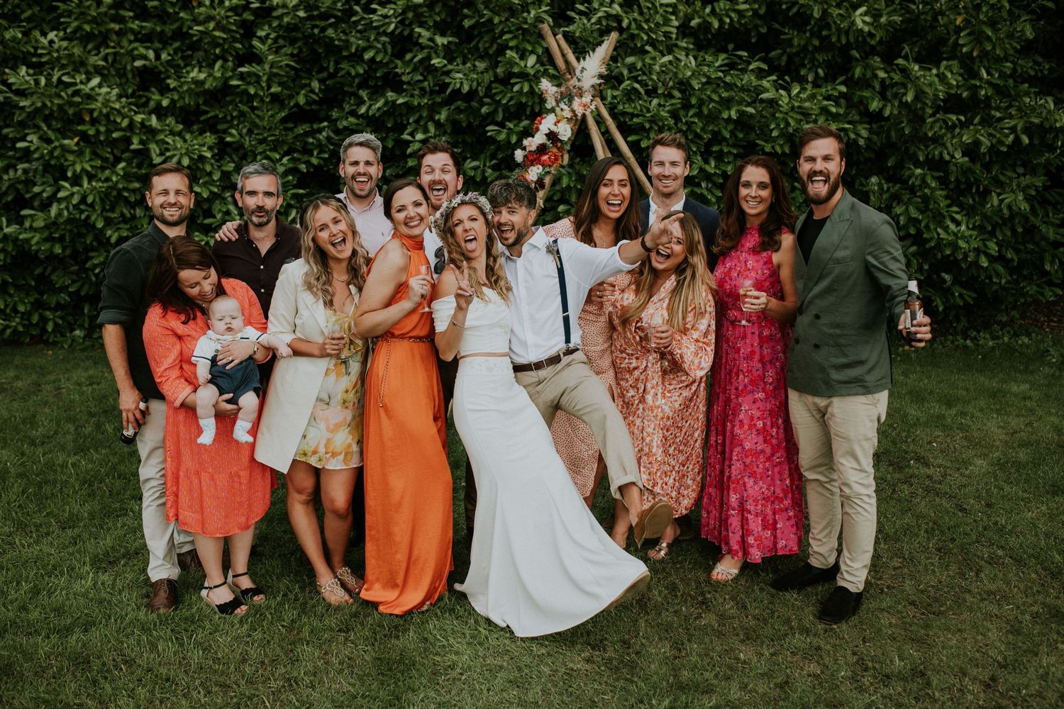 Wedding party poses joyfully on lawn with bride in white dress surrounded by colorful attire.